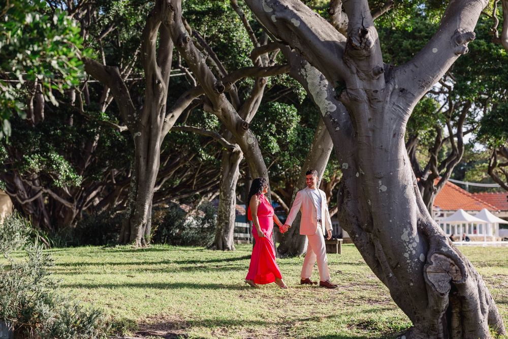 Couple holding hands walking among large trees in a lush green park, woman in red dress, man in light pink outfit.
