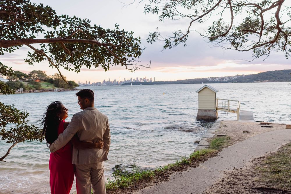 Couple embracing at Sydney Harbour waterfront at sunset with city skyline in background.