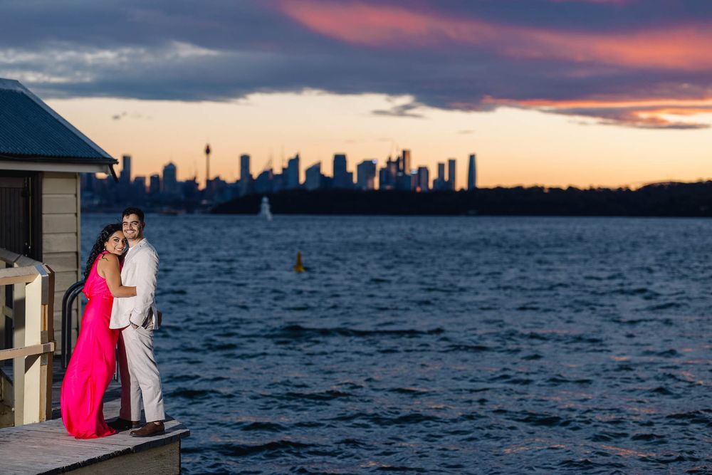 Couple posing on waterfront deck at dusk with dramatic Sydney skyline and colorful sky.