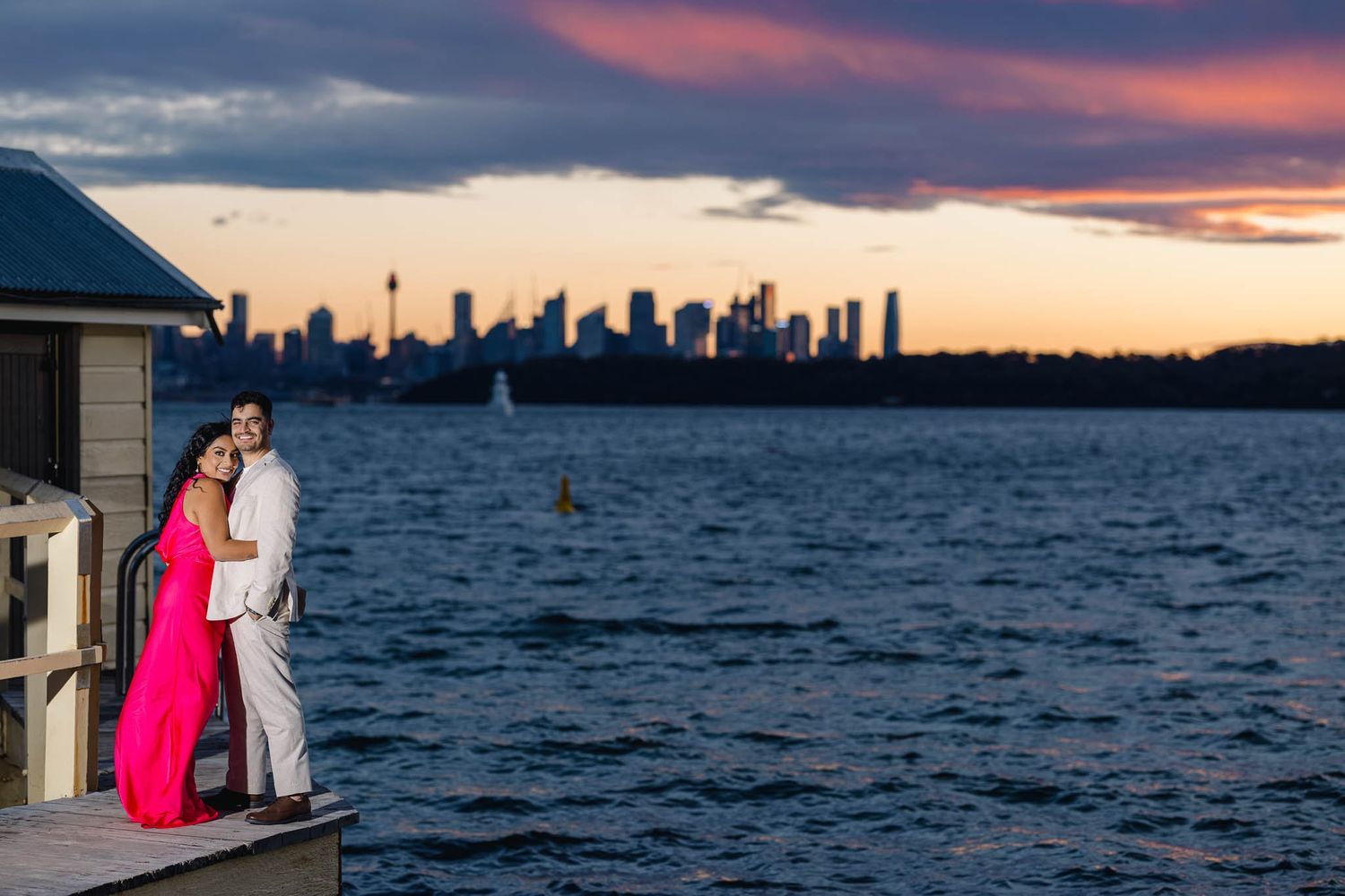 Couple posing on waterfront deck at dusk with dramatic Sydney skyline and colorful sky.