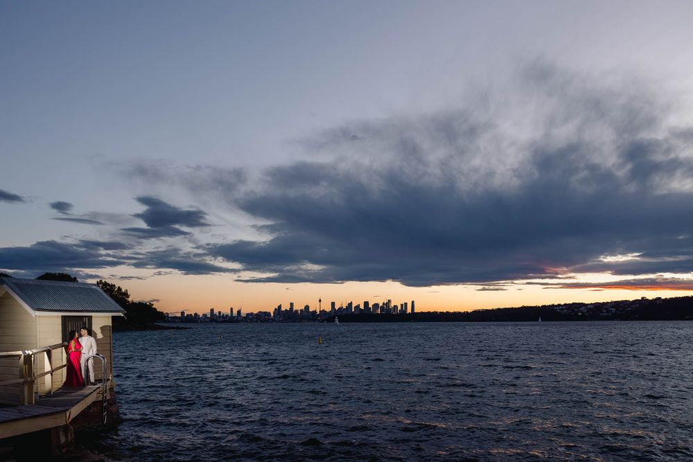 Couple on waterfront dock at sunset with Sydney city skyline silhouetted against dramatic cloudy sky.