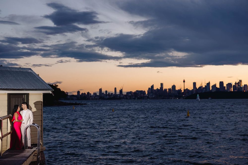 Romantic couple embracing on pier at dusk with Sydney skyline and Sydney Tower visible across harbor.