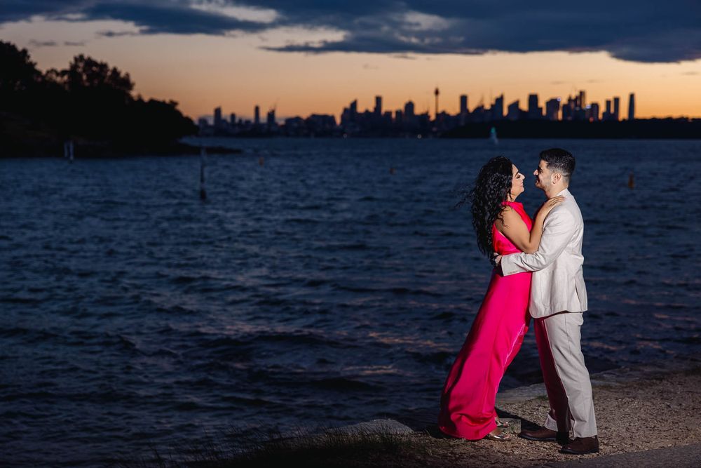 Couple embracing at sunset by Sydney Harbour with city skyline, woman in red gown, man in grey suit, romantic engagement photo.
