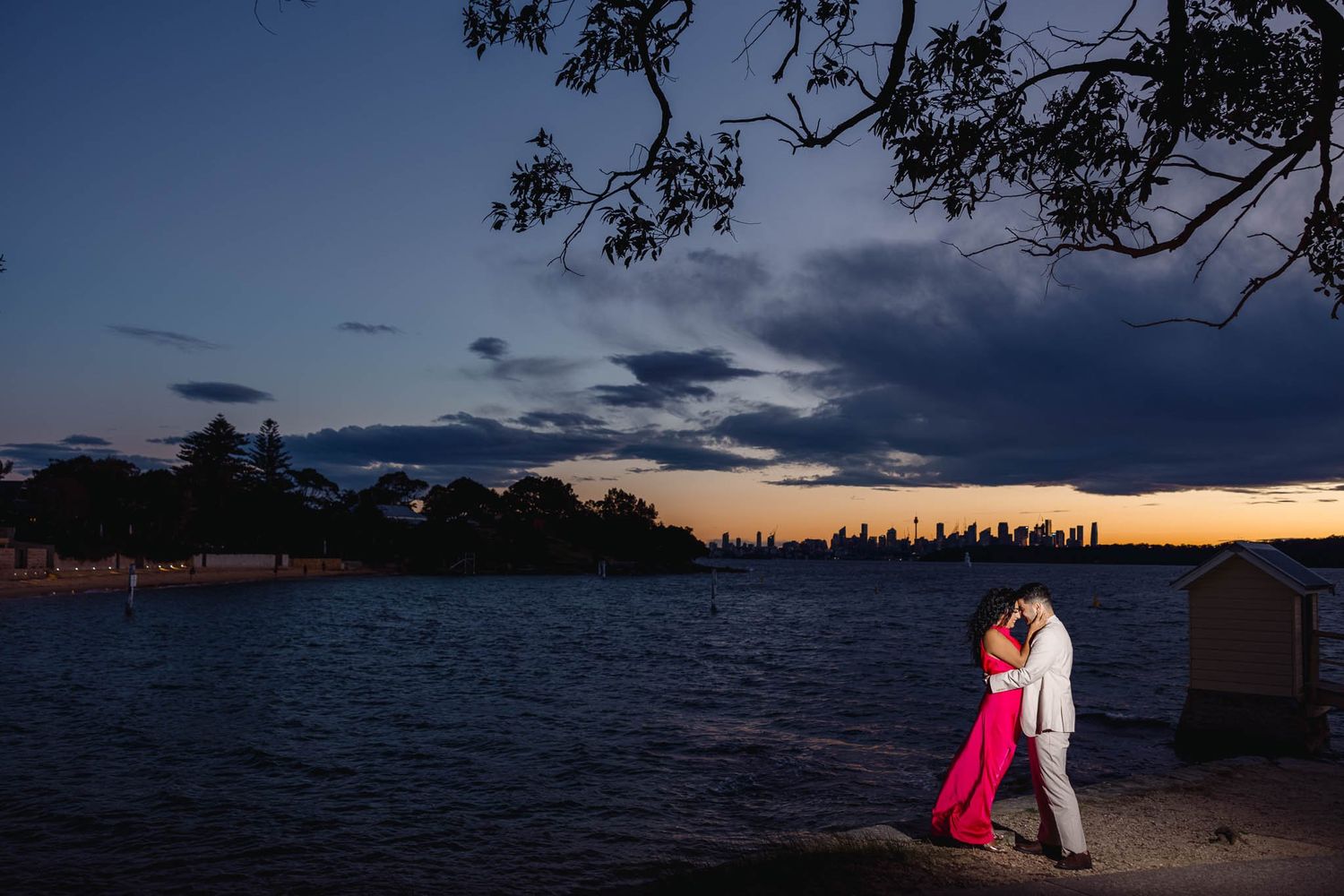 Couple embracing at sunset by Sydney Harbour waterfront, city skyline glowing in twilight, woman in pink gown.