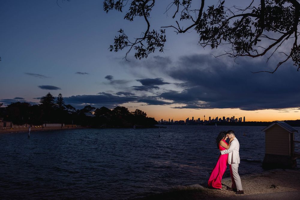 Couple embracing at sunset by Sydney Harbour waterfront, city skyline glowing in twilight, woman in pink gown.