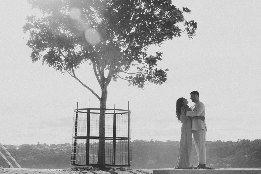 Black and white photo of a couple embracing near a tree outdoors in a romantic setting.