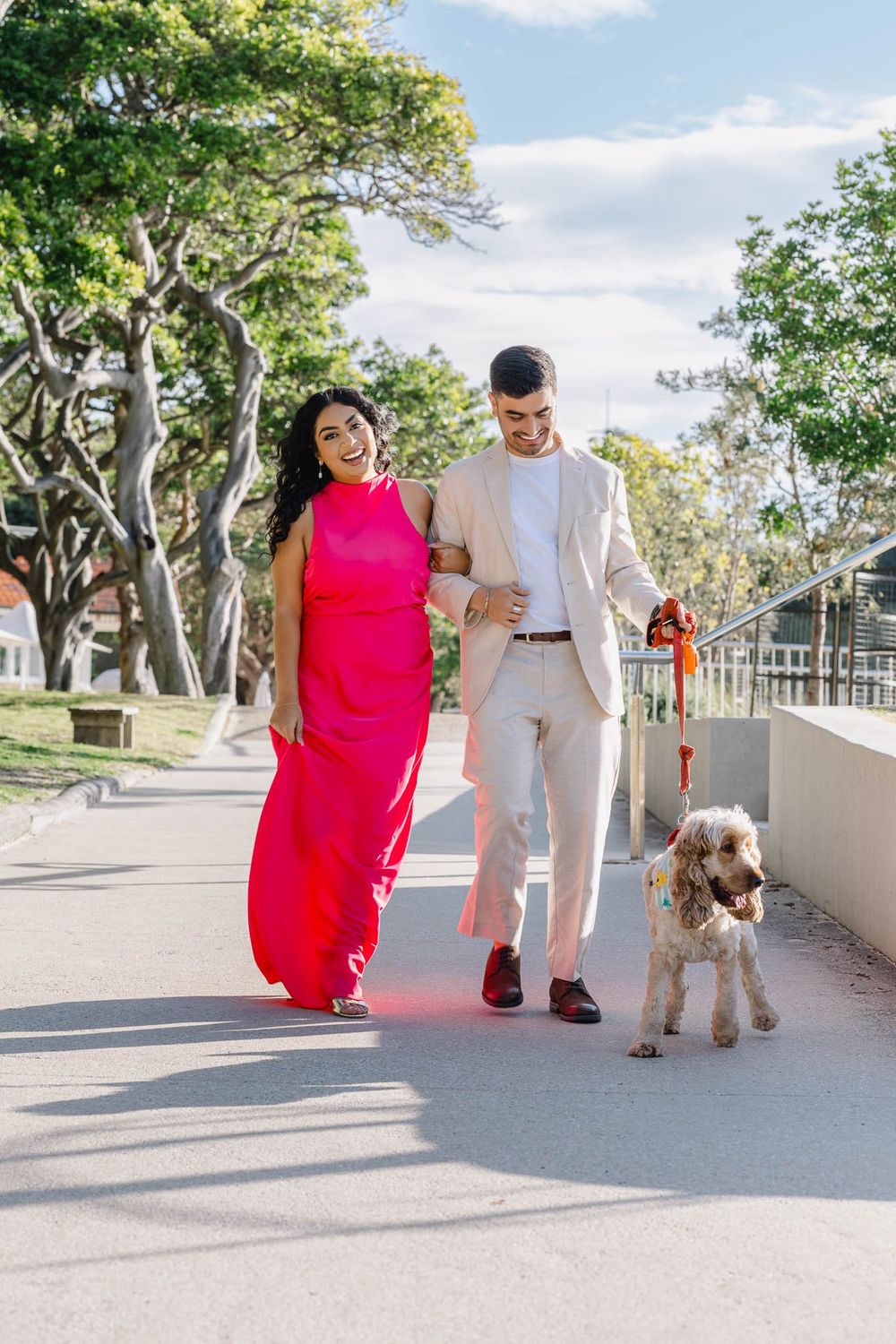 Couple in formal attire walking a fluffy dog outdoors, woman in pink dress, man in beige suit, tree-lined path.