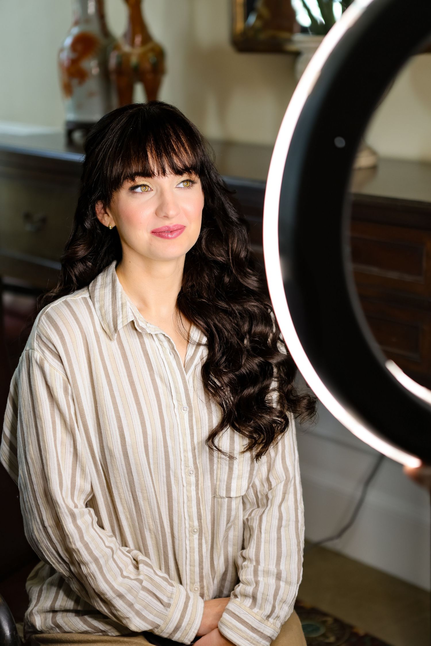 Young woman with dark curly hair and bangs poses near a ring light in a striped shirt indoors.