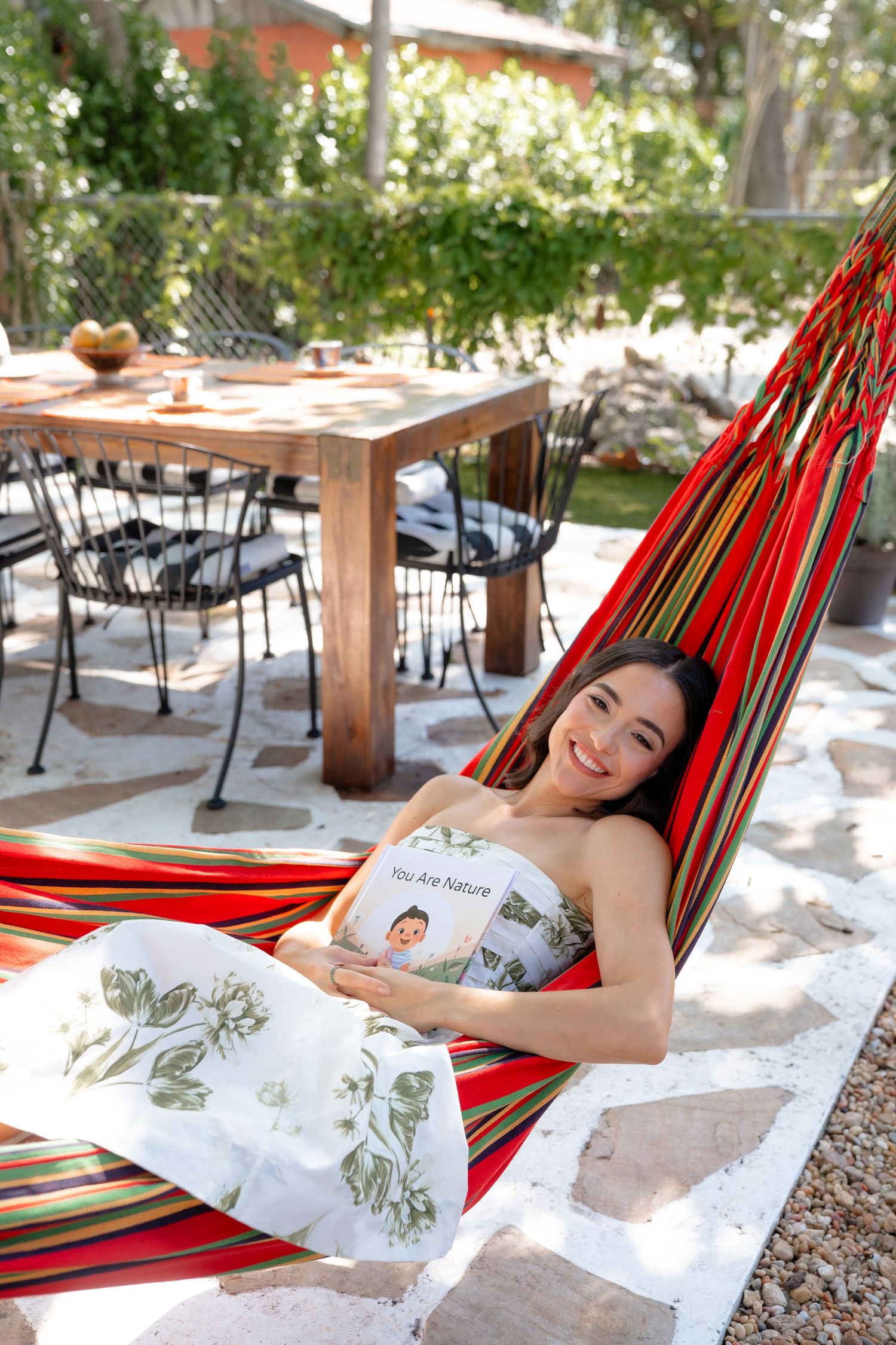 Woman relaxing in a colorful striped hammock outdoors, holding a book titled You Are Nature, smiling in a garden setting.