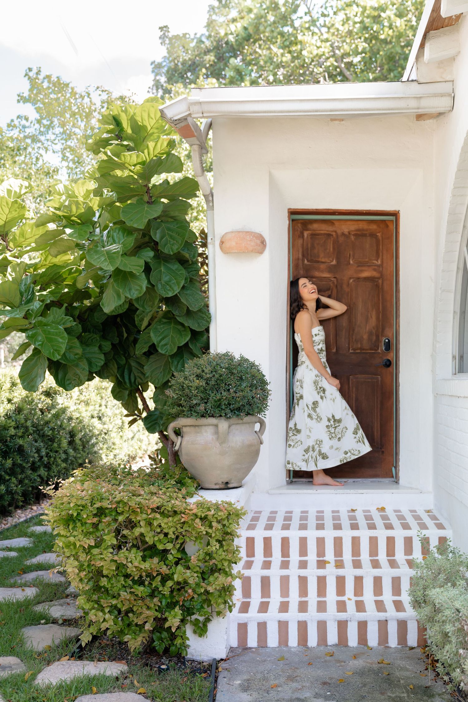 Woman in floral dress standing at wooden front door of white stucco home with lush garden and brick steps.