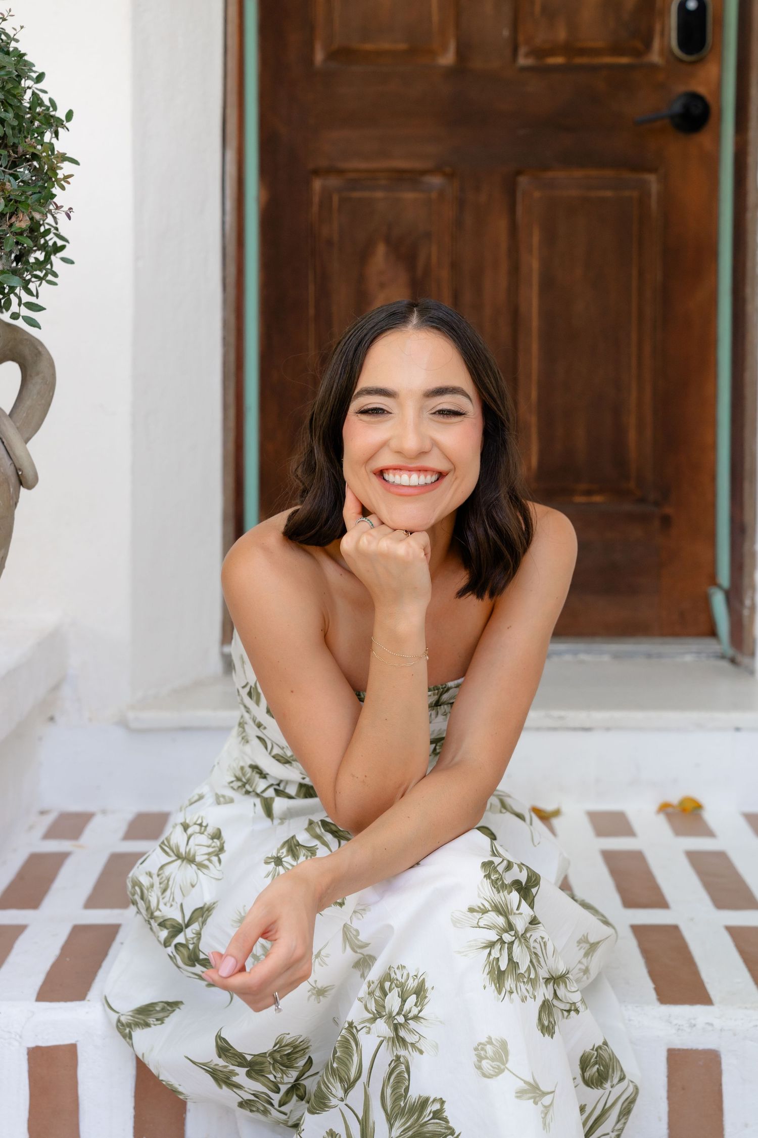 Smiling woman in floral dress sitting on white steps in front of a wooden door.