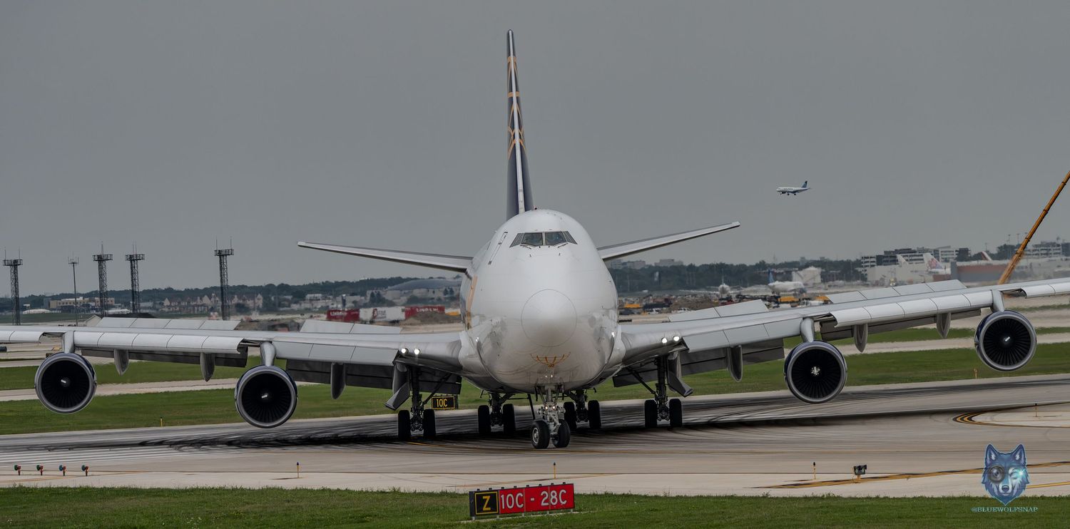 Large white commercial Boeing 747 aircraft taxiing on runway at busy airport under overcast skies.