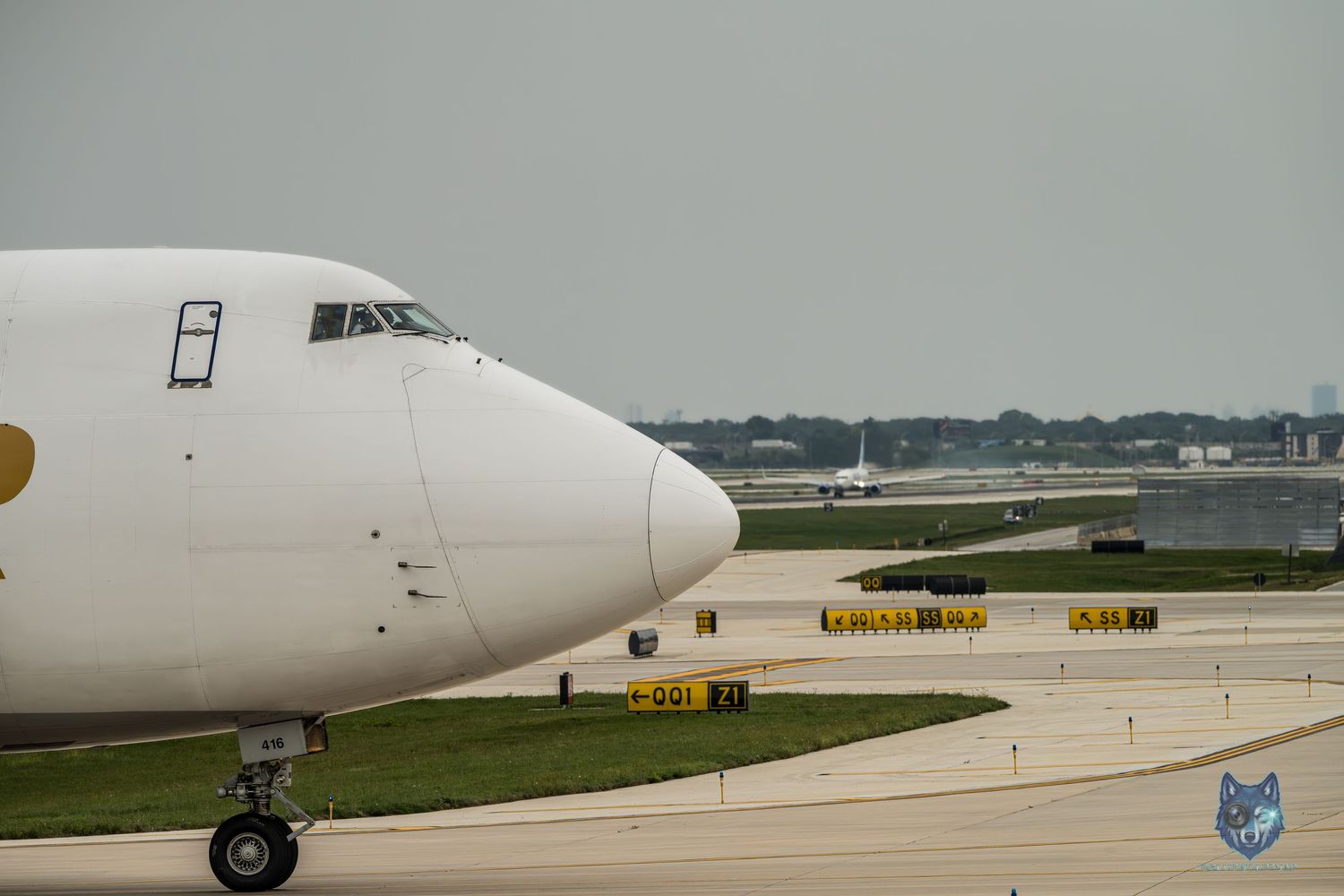 White cargo aircraft taxiing on airport runway with yellow taxiway signs visible in the background on a cloudy day.