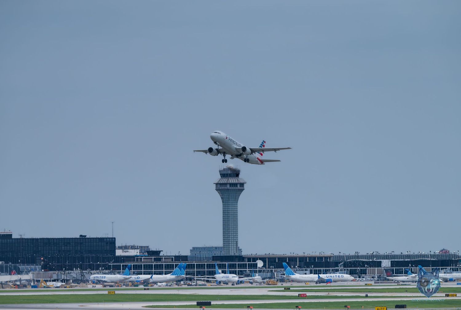Airplane taking off near airport control tower with multiple aircraft parked at terminal gates under overcast sky.