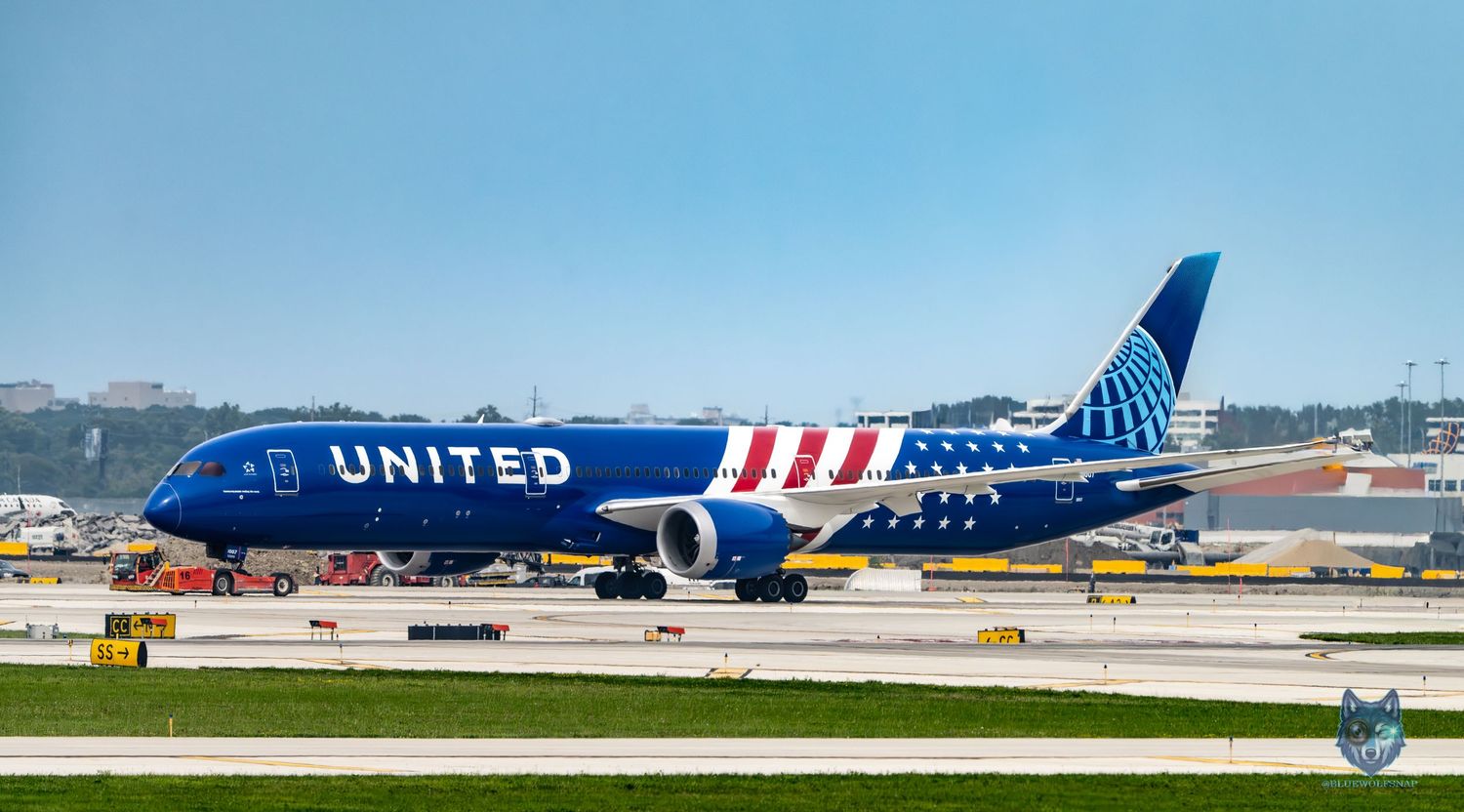 United Airlines Boeing 757 in blue livery taxiing on runway at busy airport on a clear sunny day.