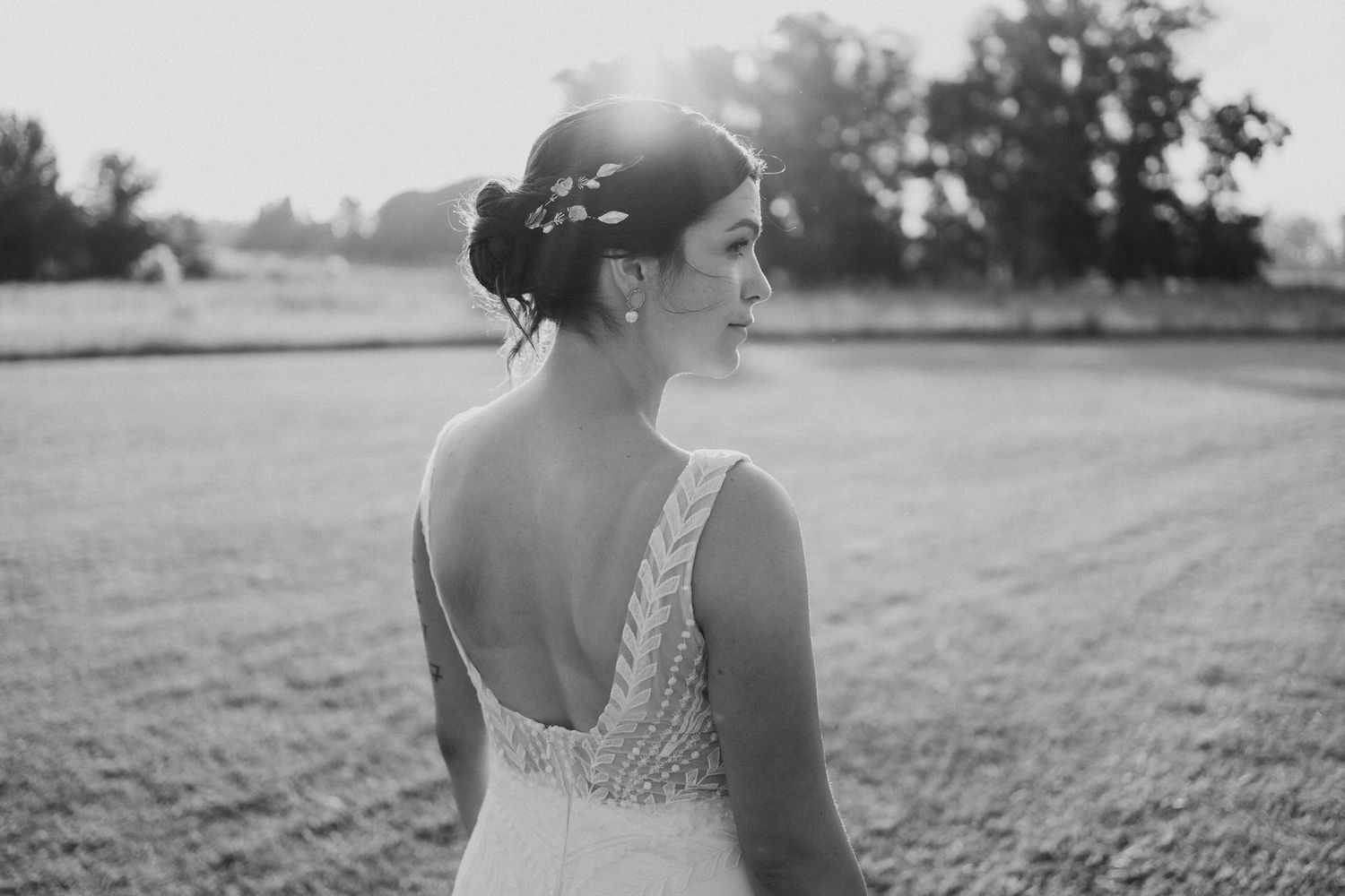 Bride with elegant updo and low-back beaded wedding gown standing in sunlit field, black and white portrait.
