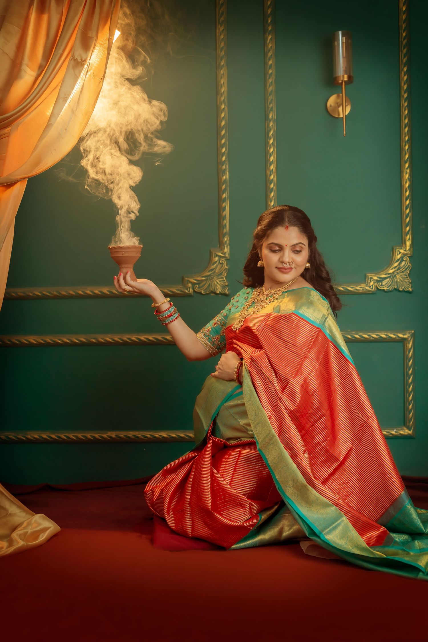 Expecting mother in a red and green silk saree kneeling beside a smoking clay pot against an ornate emerald green backdrop