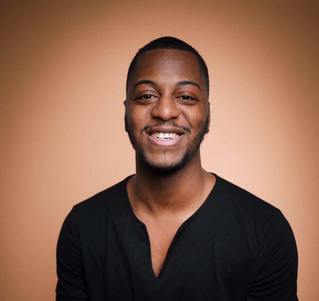 Smiling young Black man in black v-neck shirt poses against warm brown background in professional portrait photo.