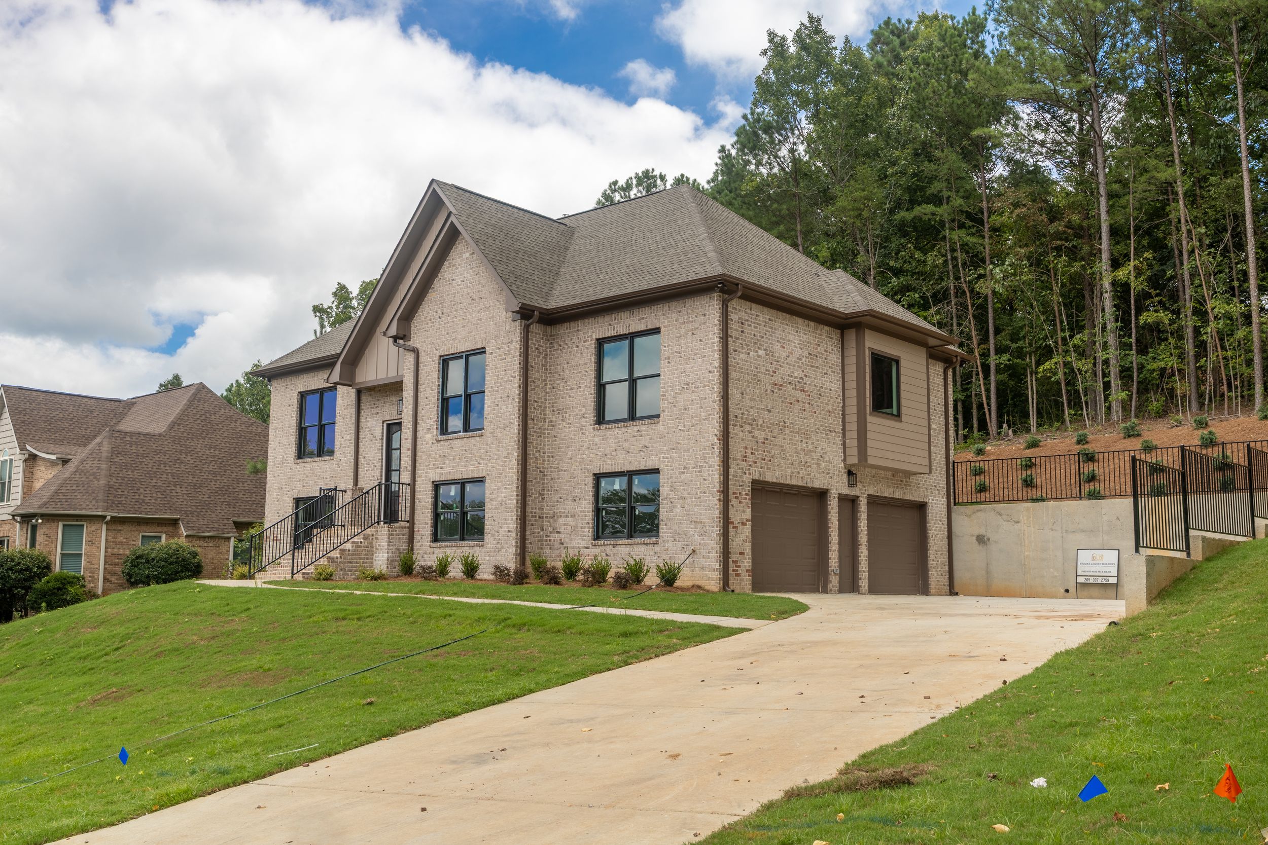 New brick two-story home with attached garage, manicured lawn, and wooded backdrop on a cloudy day.