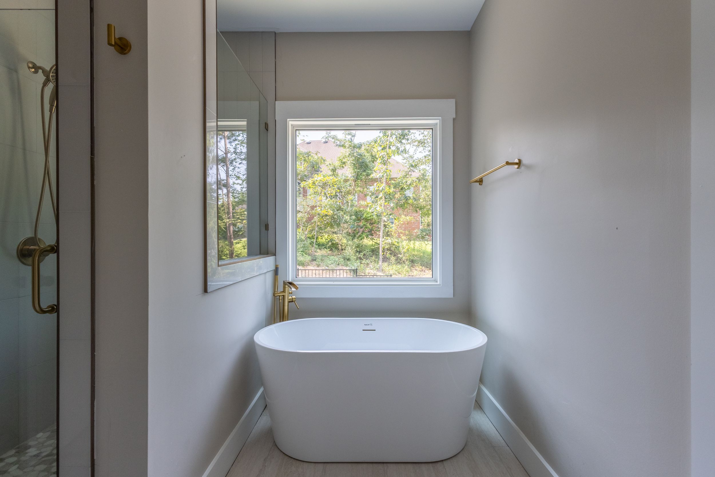 Modern bathroom with freestanding white soaking tub, brass fixtures, and large window with garden view.