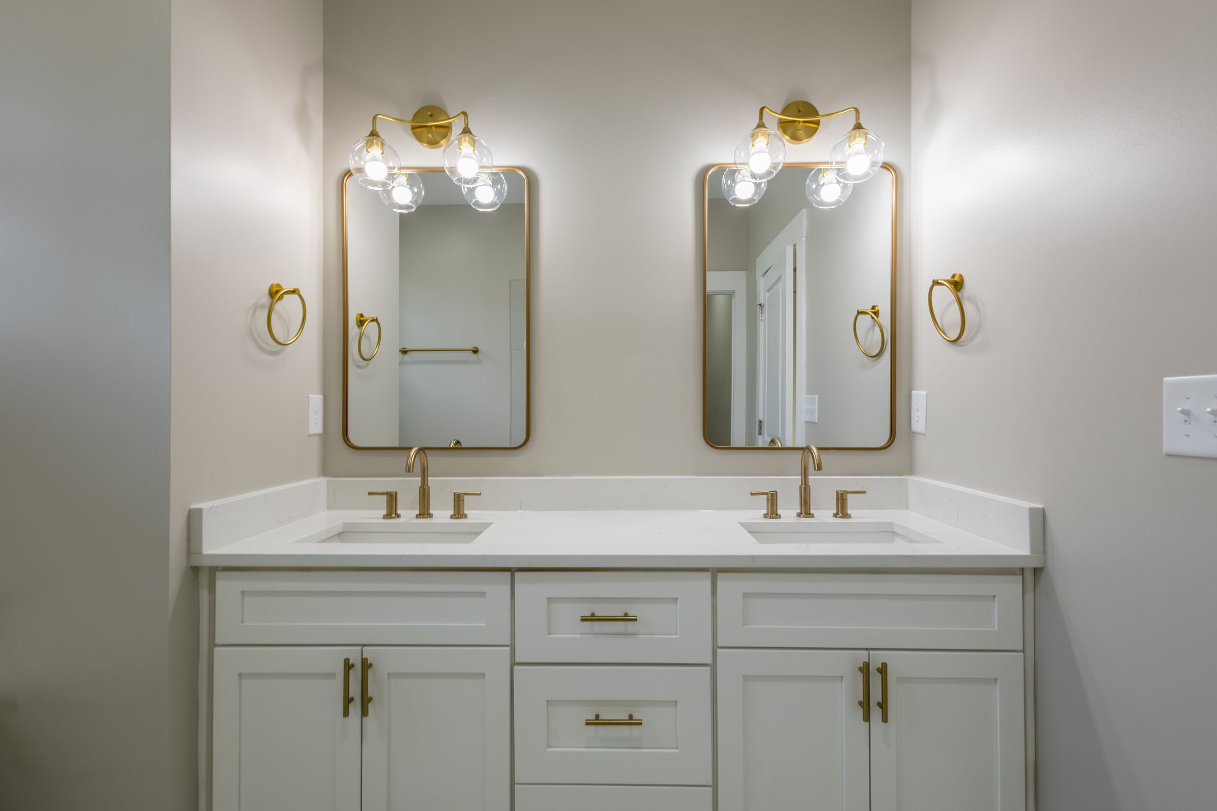 Double vanity bathroom with white cabinets, gold hardware, dual mirrors, and warm brass light fixtures on neutral walls.