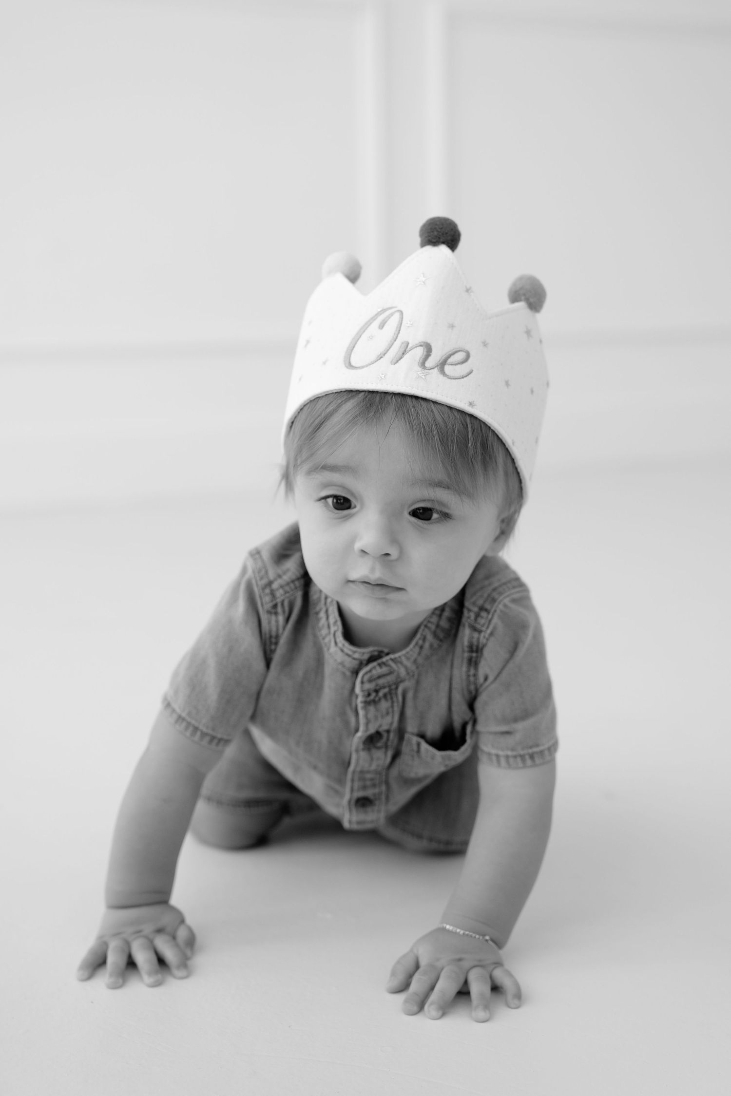 Black and white photo of a baby wearing a crown hat that says One, crawling on the floor during first birthday.