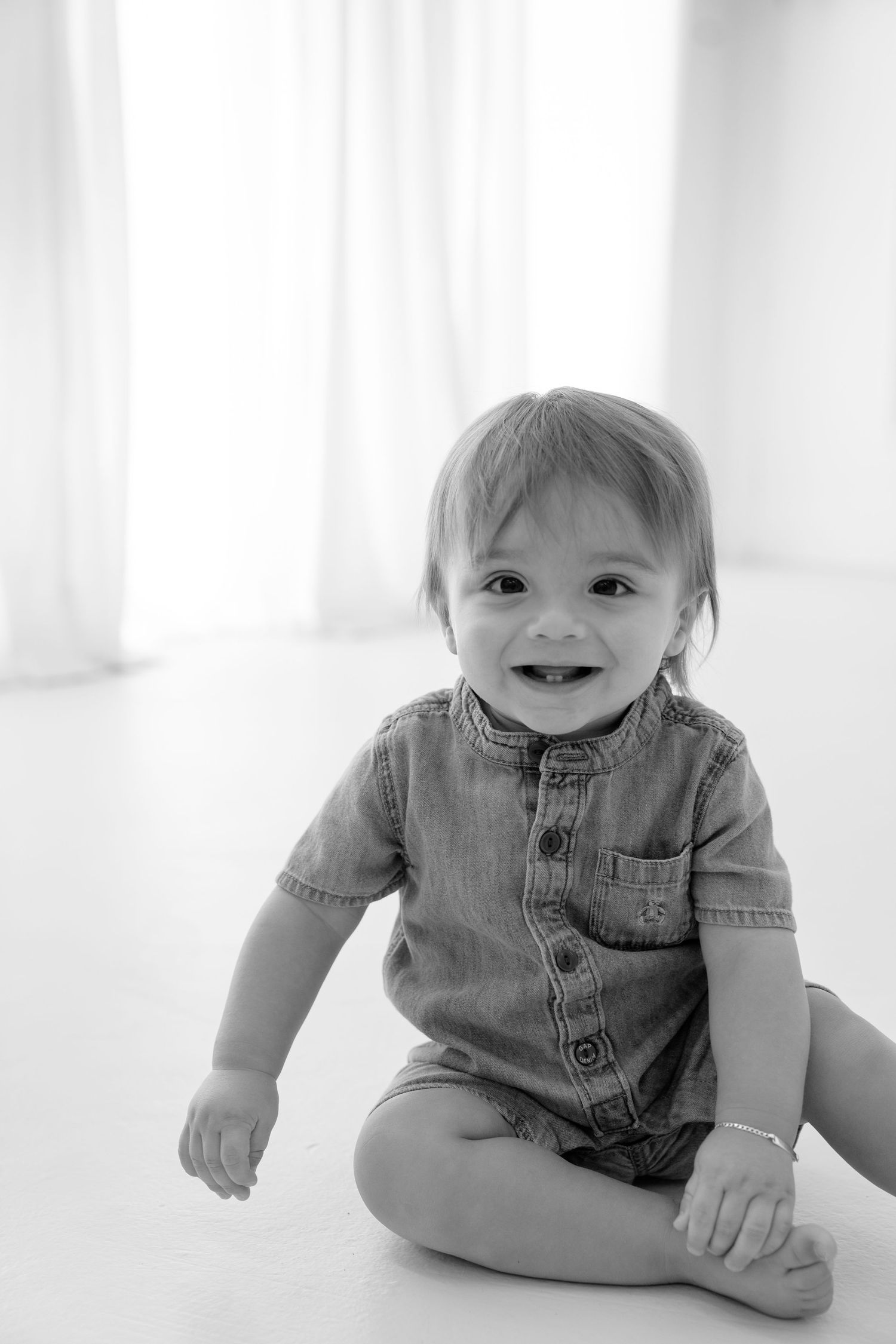 Black and white photo of a smiling baby boy sitting on floor wearing a denim romper.
