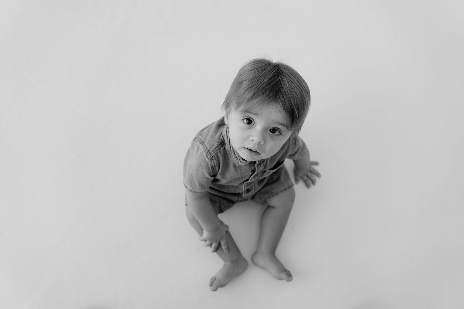 Black and white photo of a curious baby sitting on a white background, looking up at the camera.