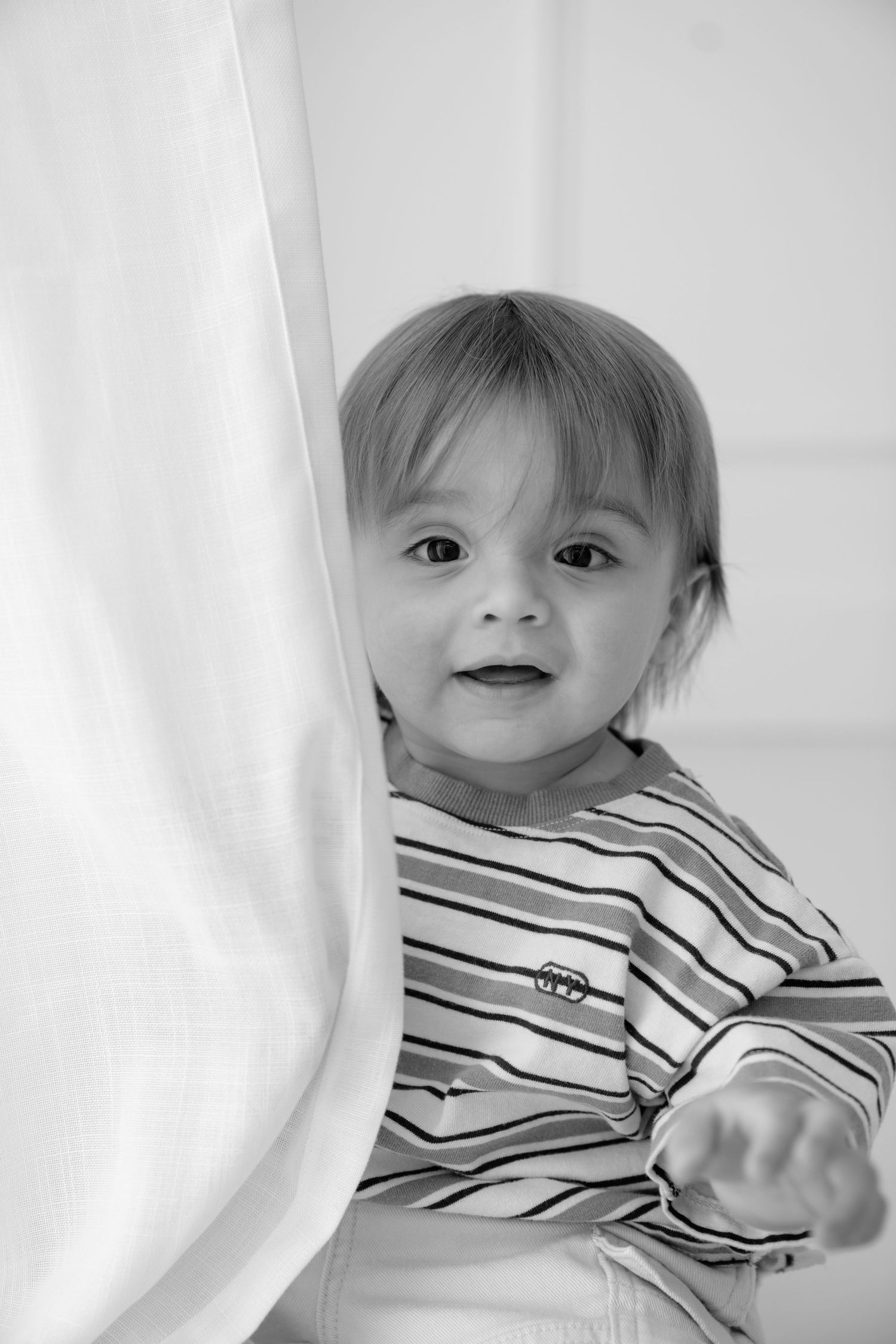 Black and white photo of a cute toddler peeking around a white curtain wearing a striped shirt.