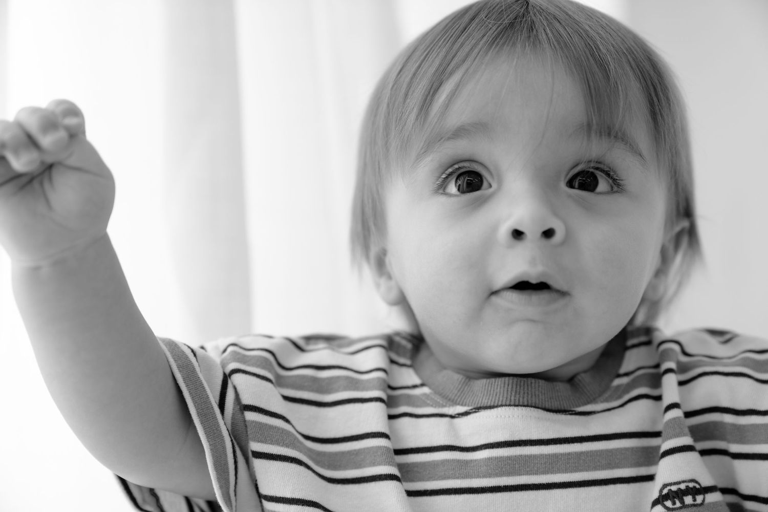Black and white photo of a curious baby with wide eyes raising one fist, wearing a striped shirt.