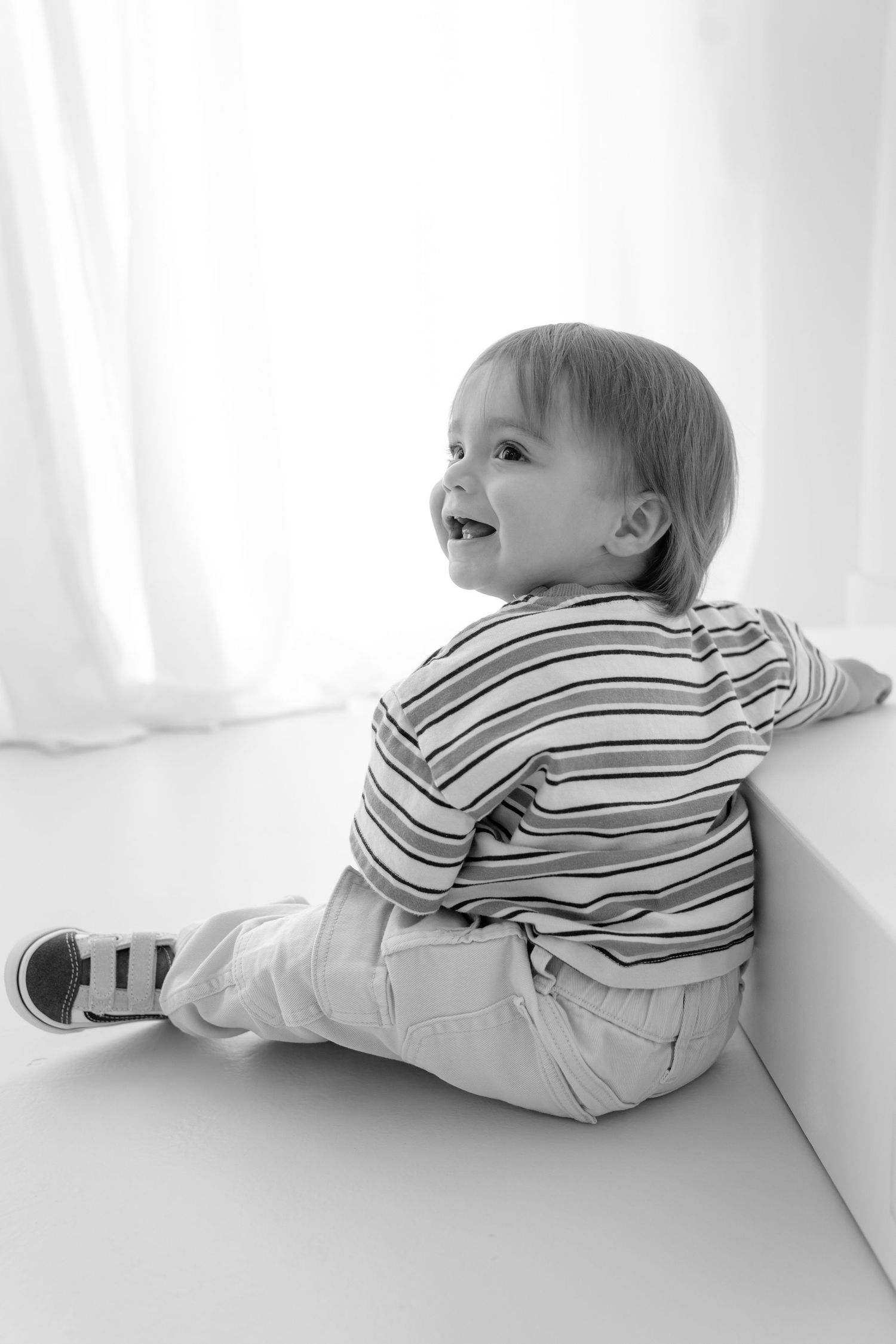 Black and white photo of a happy toddler boy sitting on floor in striped shirt and sneakers, smiling and looking up.