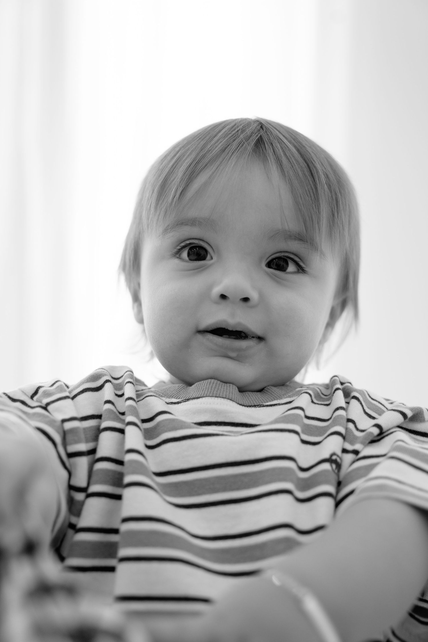 Black and white portrait of an adorable baby with wide eyes wearing a striped shirt, looking curiously at the camera.