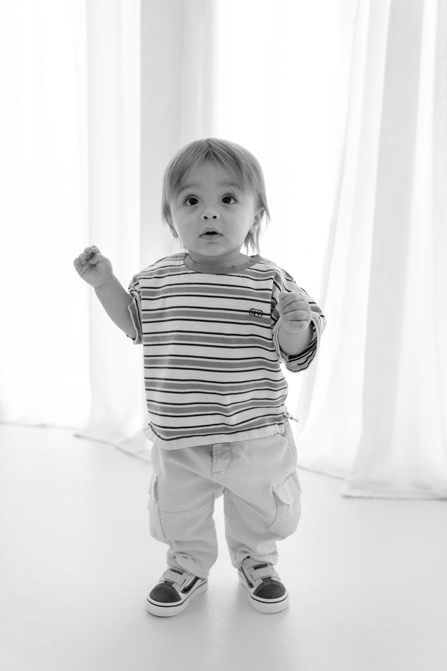 Black and white photo of a curious toddler boy in striped shirt and cargo pants standing with fist raised.
