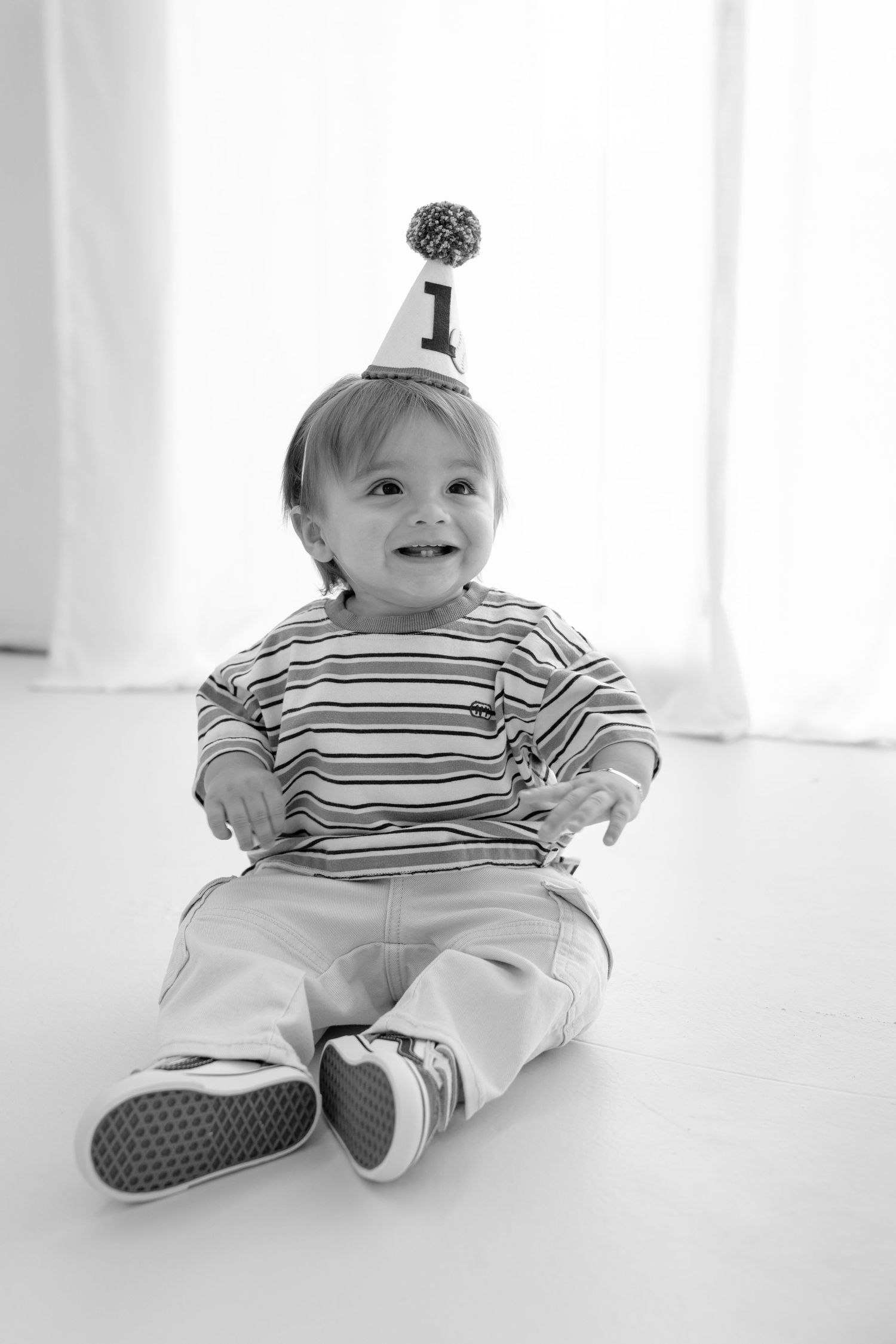 Black and white photo of a smiling baby wearing a number 1 birthday hat and striped shirt, sitting on floor.