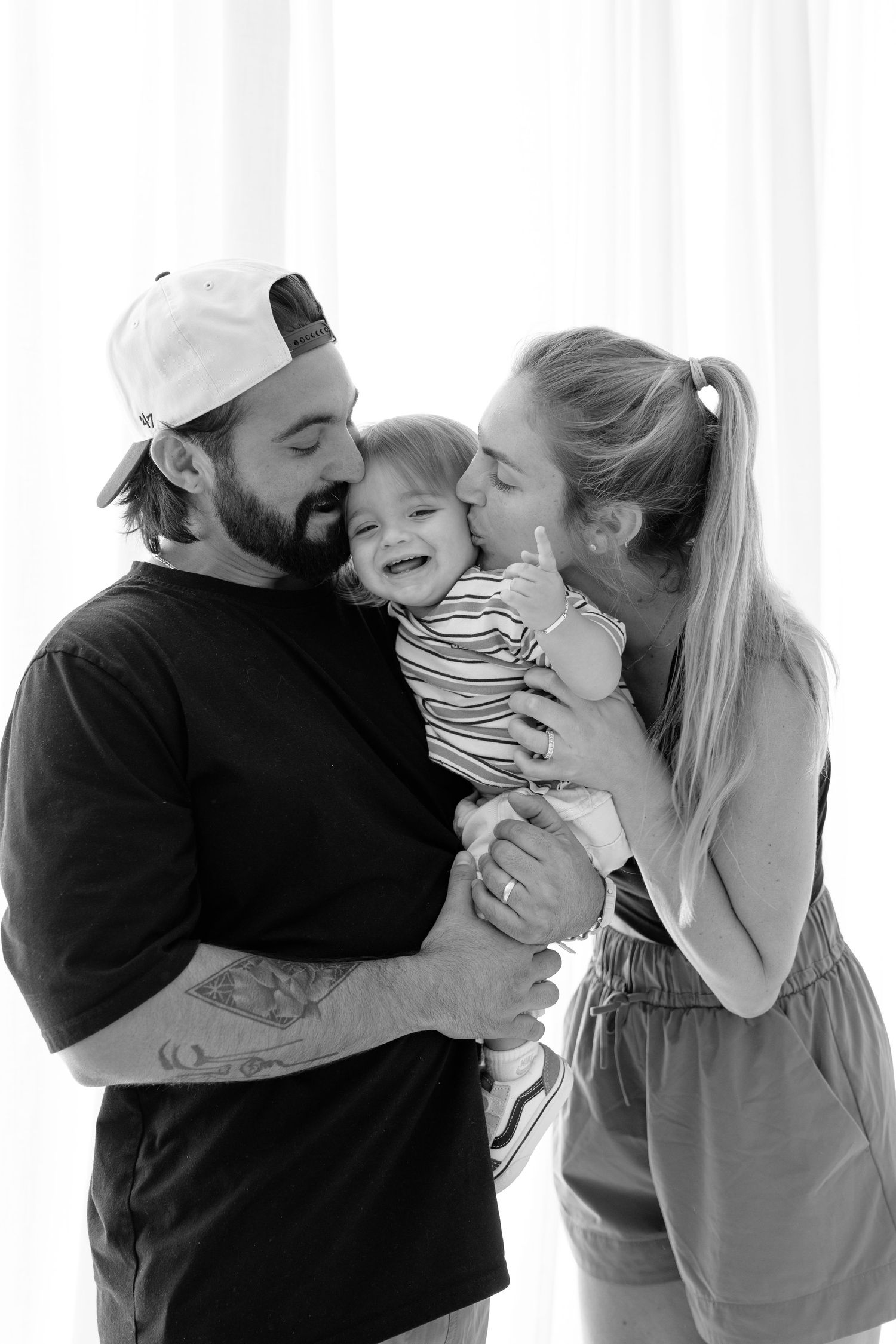 Black and white photo of a bearded dad in a cap and tattooed arms holding a smiling baby while mom kisses the child.