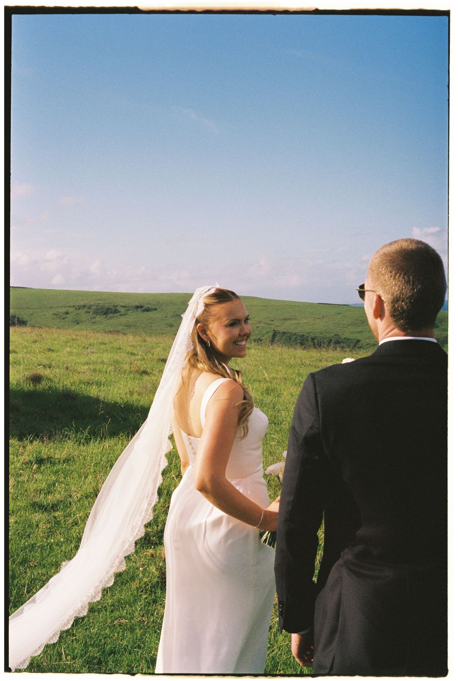 Smiling bride in white gown and lace veil holding hands with groom in black suit in green field.
