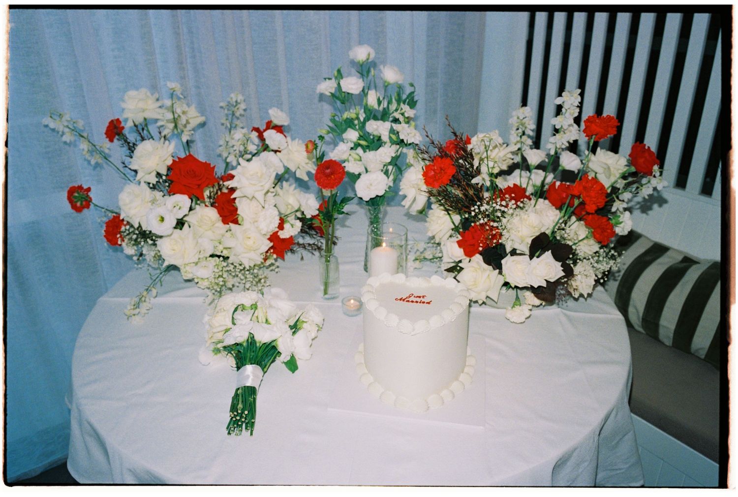 White wedding cake surrounded by red and white floral arrangements on a white tablecloth-covered round table.