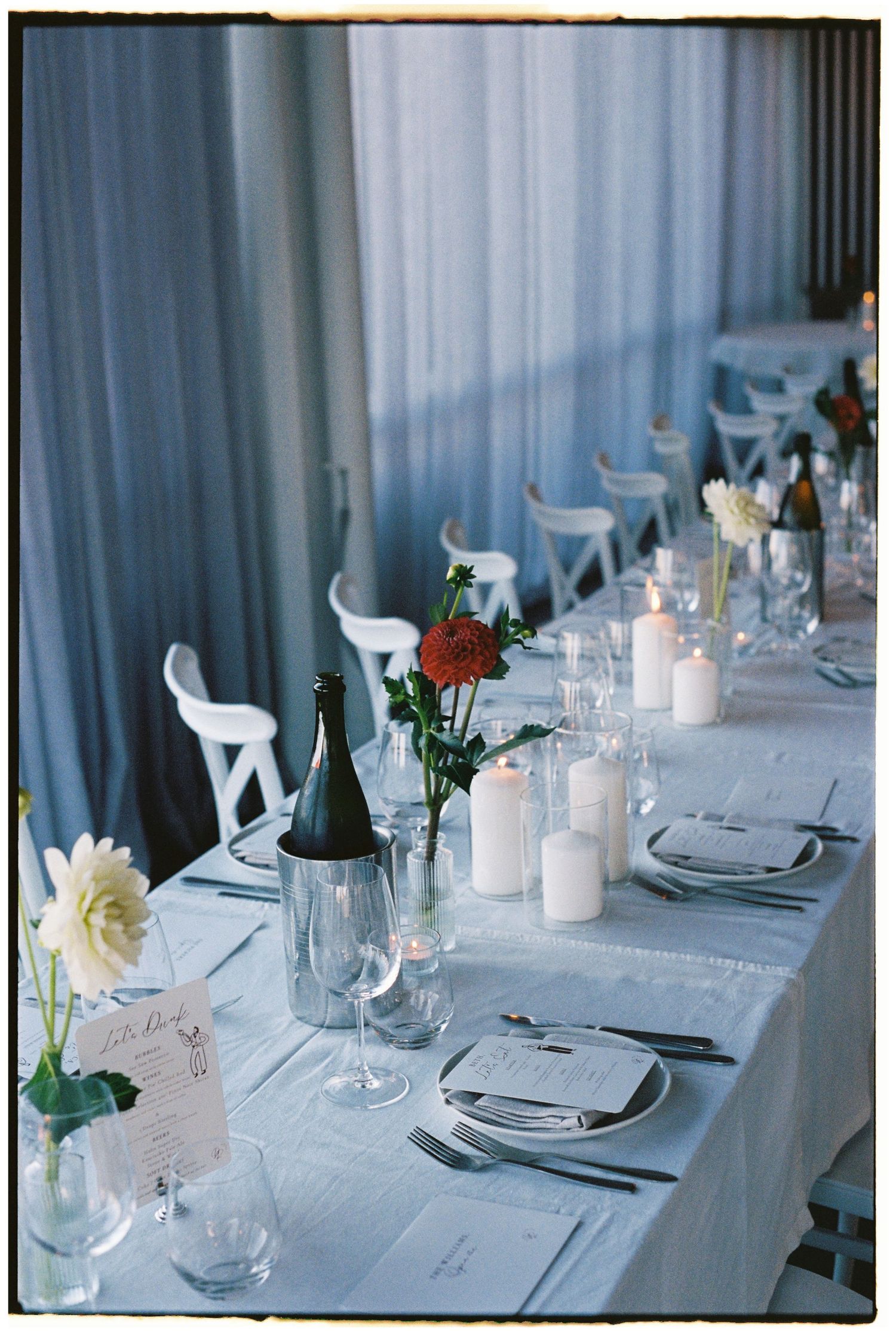 Elegant wedding reception table setting with white linens, candles, red roses, and wine bottles in soft blue lighting.
