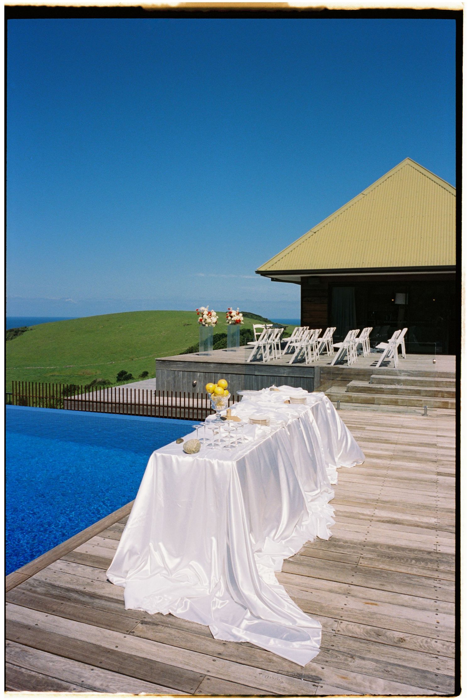 Elegant white draped wedding reception table on wooden deck beside infinity pool with ocean and green hills backdrop.