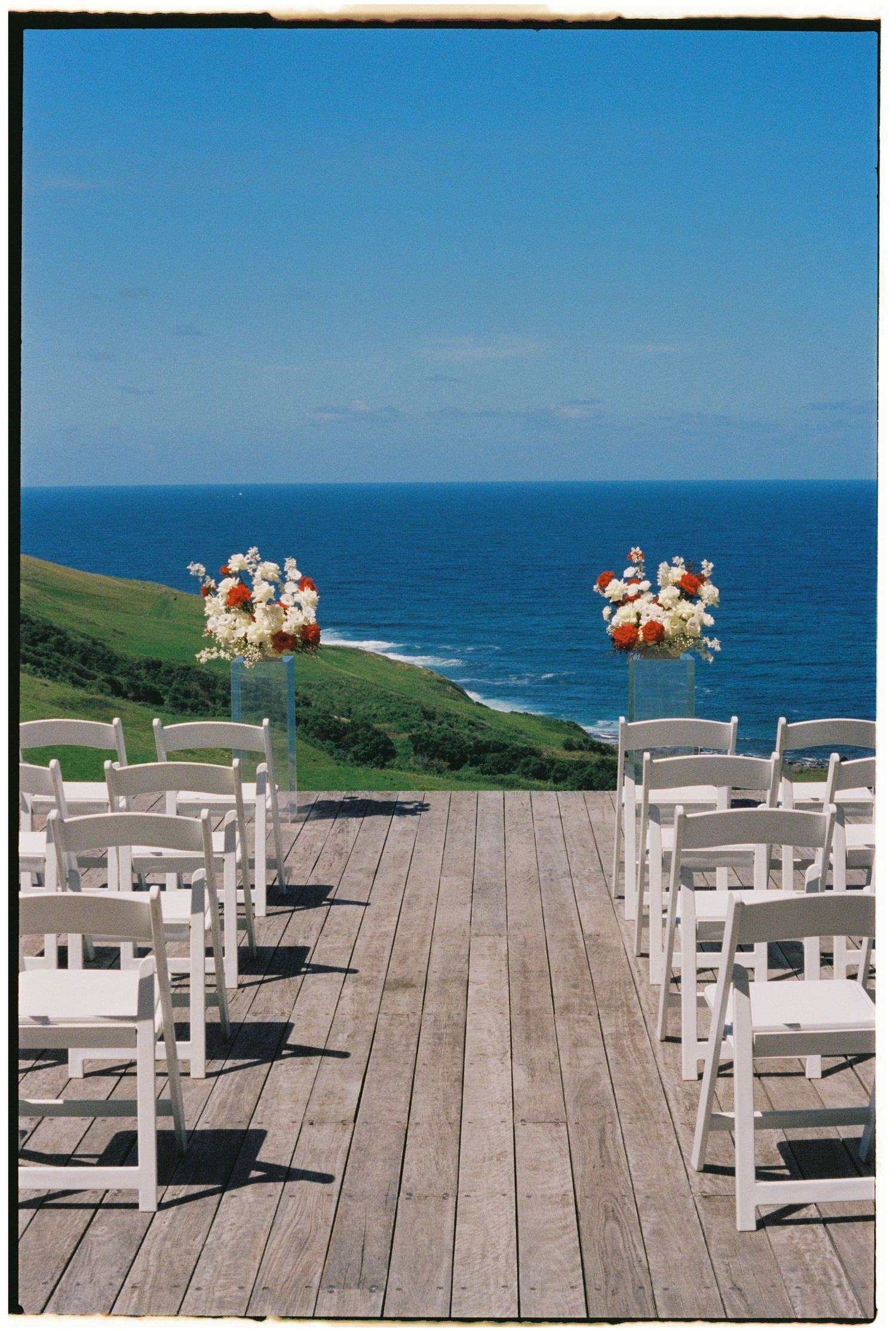 Outdoor wedding ceremony setup with white chairs and floral arrangements on wooden deck overlooking scenic ocean coastline.