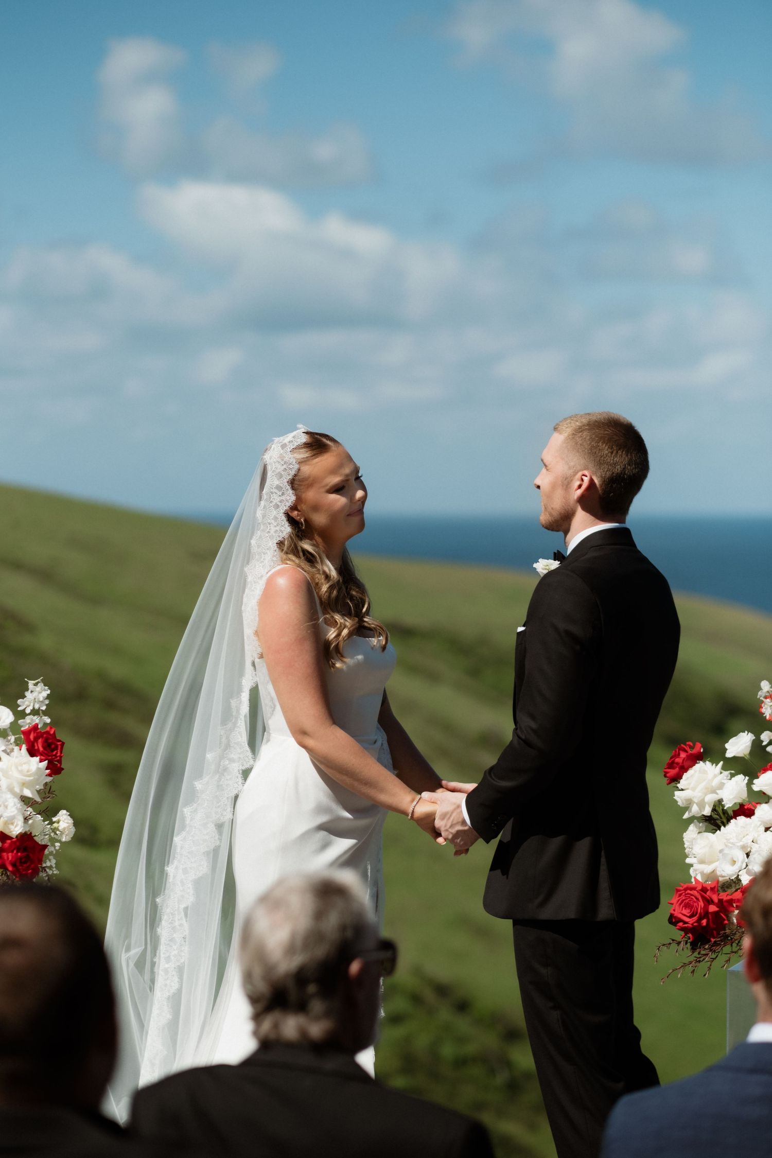 A bride and groom hold hands at an outdoor ceremony on a green hillside with a blue ocean backdrop.