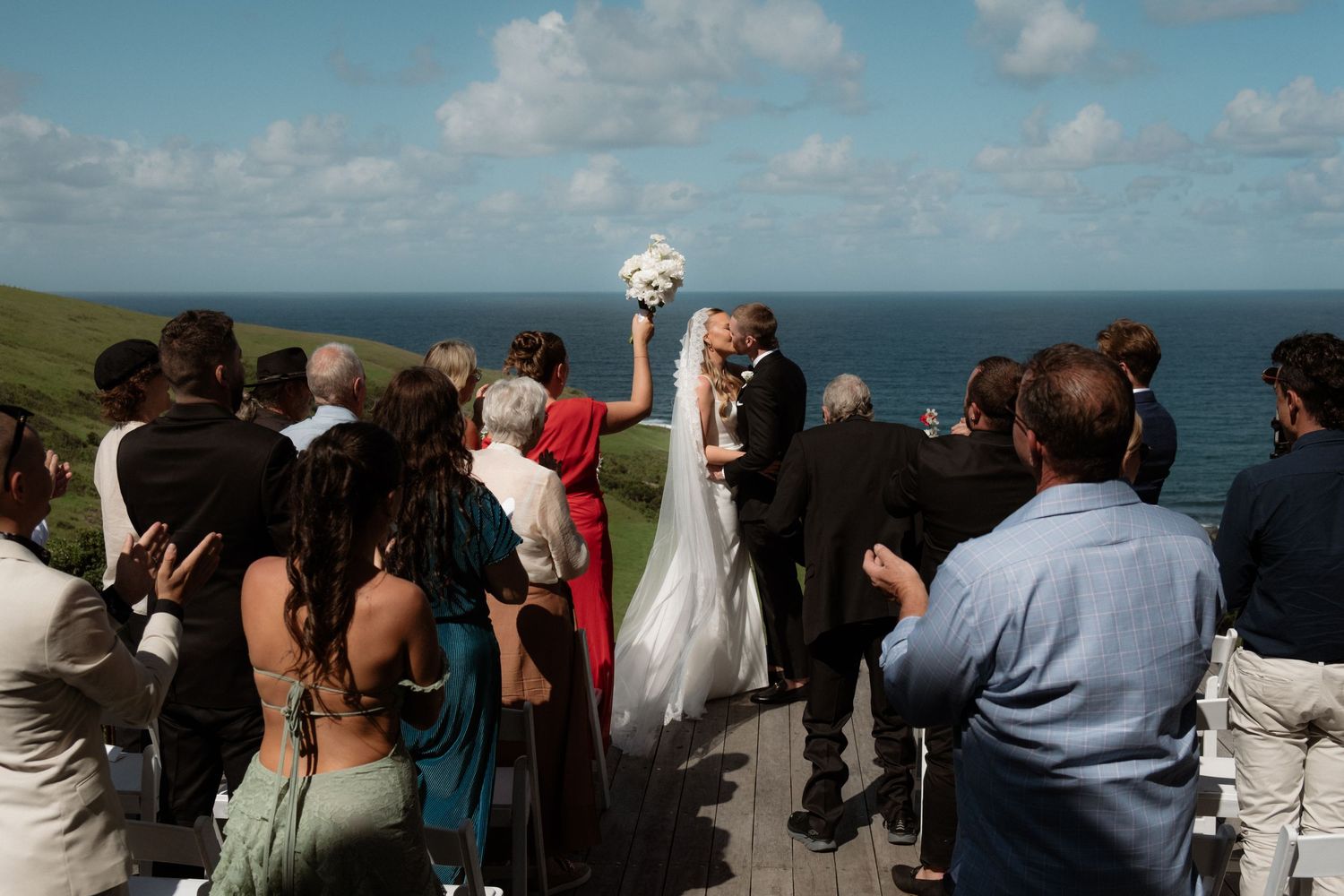Bride and groom kiss at outdoor coastal wedding ceremony on wooden deck with ocean views and guests watching.