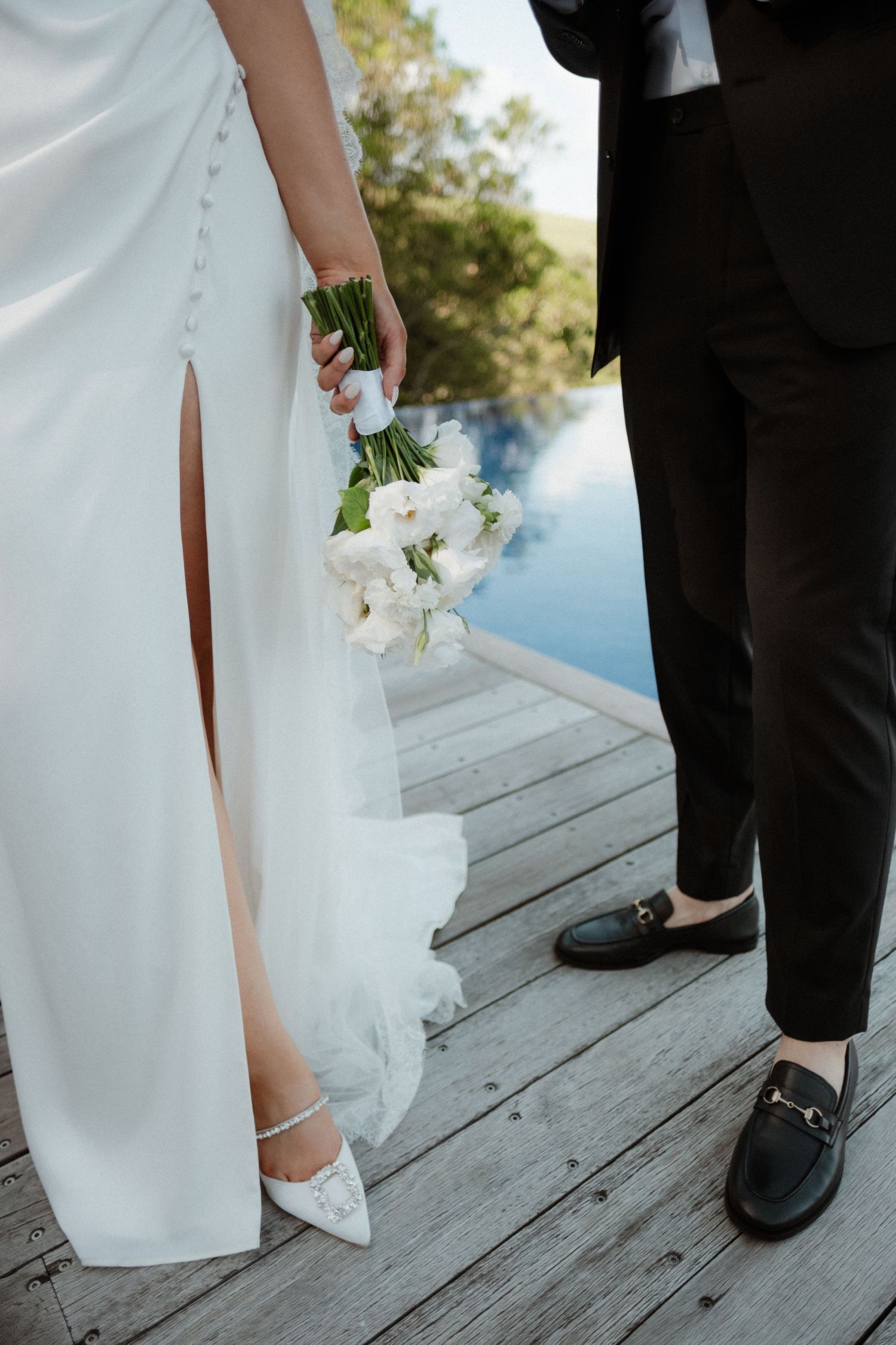 A bride in a white gown holding a small white floral bouquet stands beside a groom in black dress shoes.