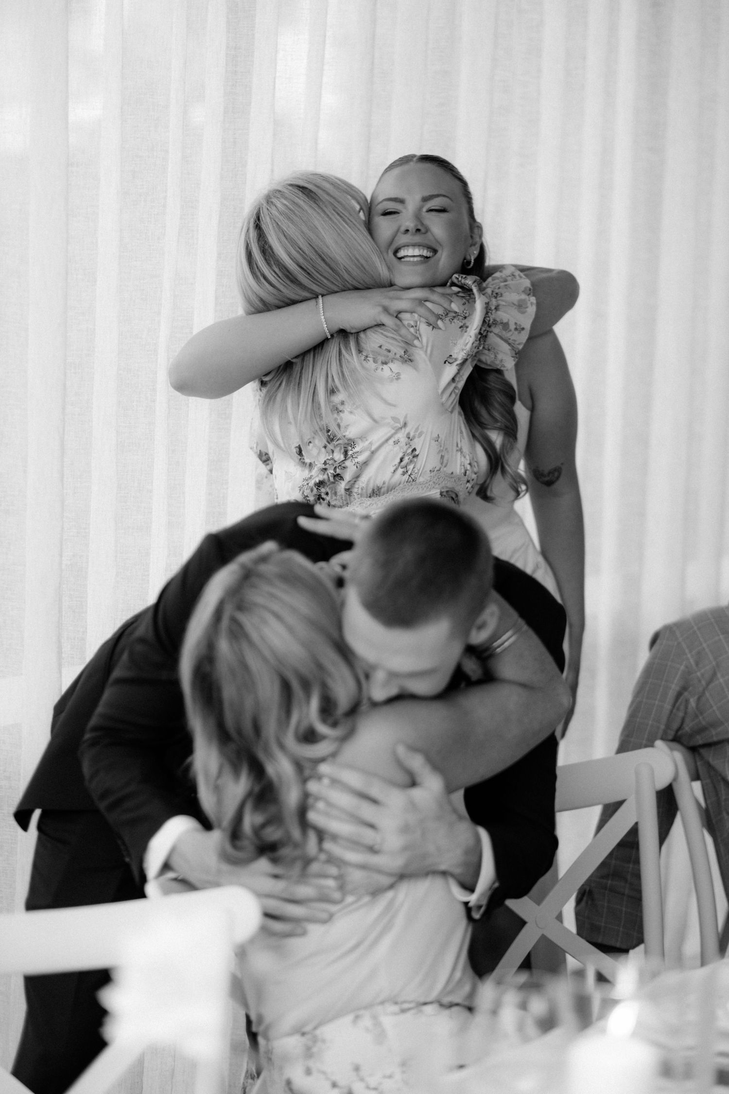 Black and white photo of joyful wedding guests sharing warm embraces at a reception venue.
