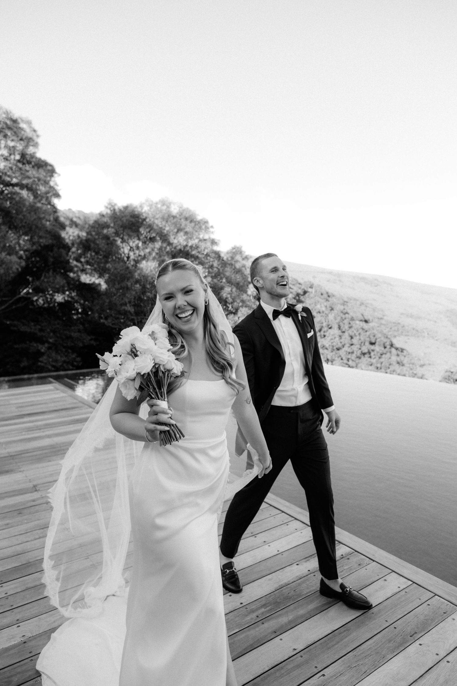 Smiling bride in white gown with veil holds bouquet as groom laughs behind her on wooden deck.