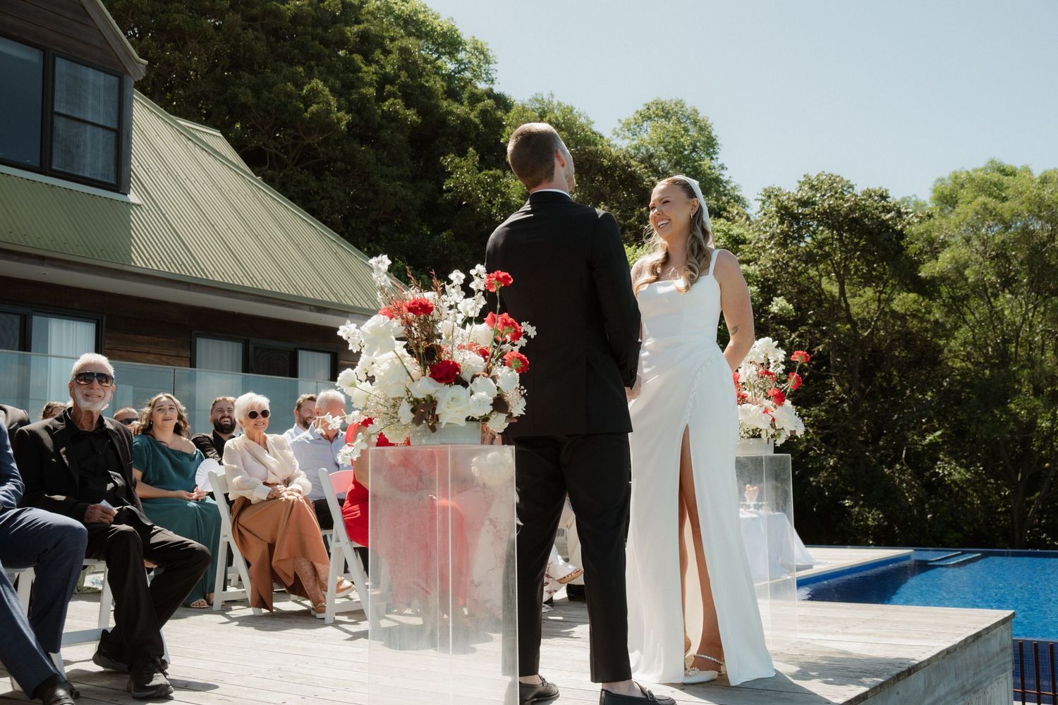 Bride and groom exchange vows at an outdoor poolside wedding ceremony with red and white floral arrangements.