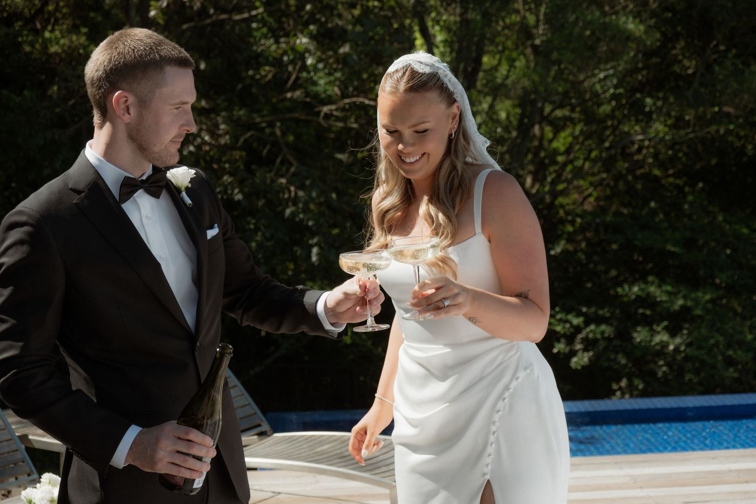 Bride and groom toasting with champagne coupes beside a pool at their outdoor wedding reception.