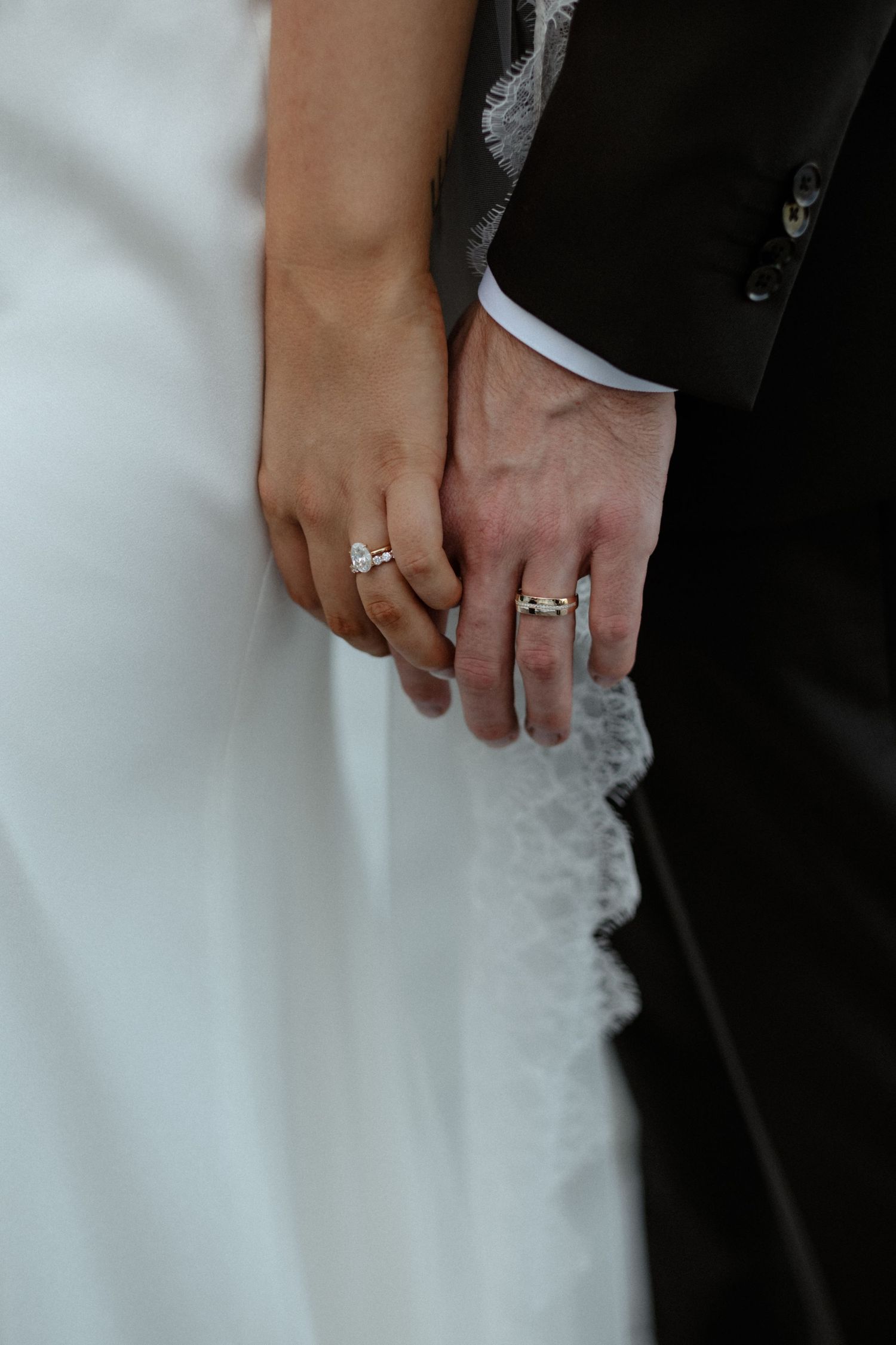 Bride and groom holding hands showing wedding rings, wearing white gown and black suit.