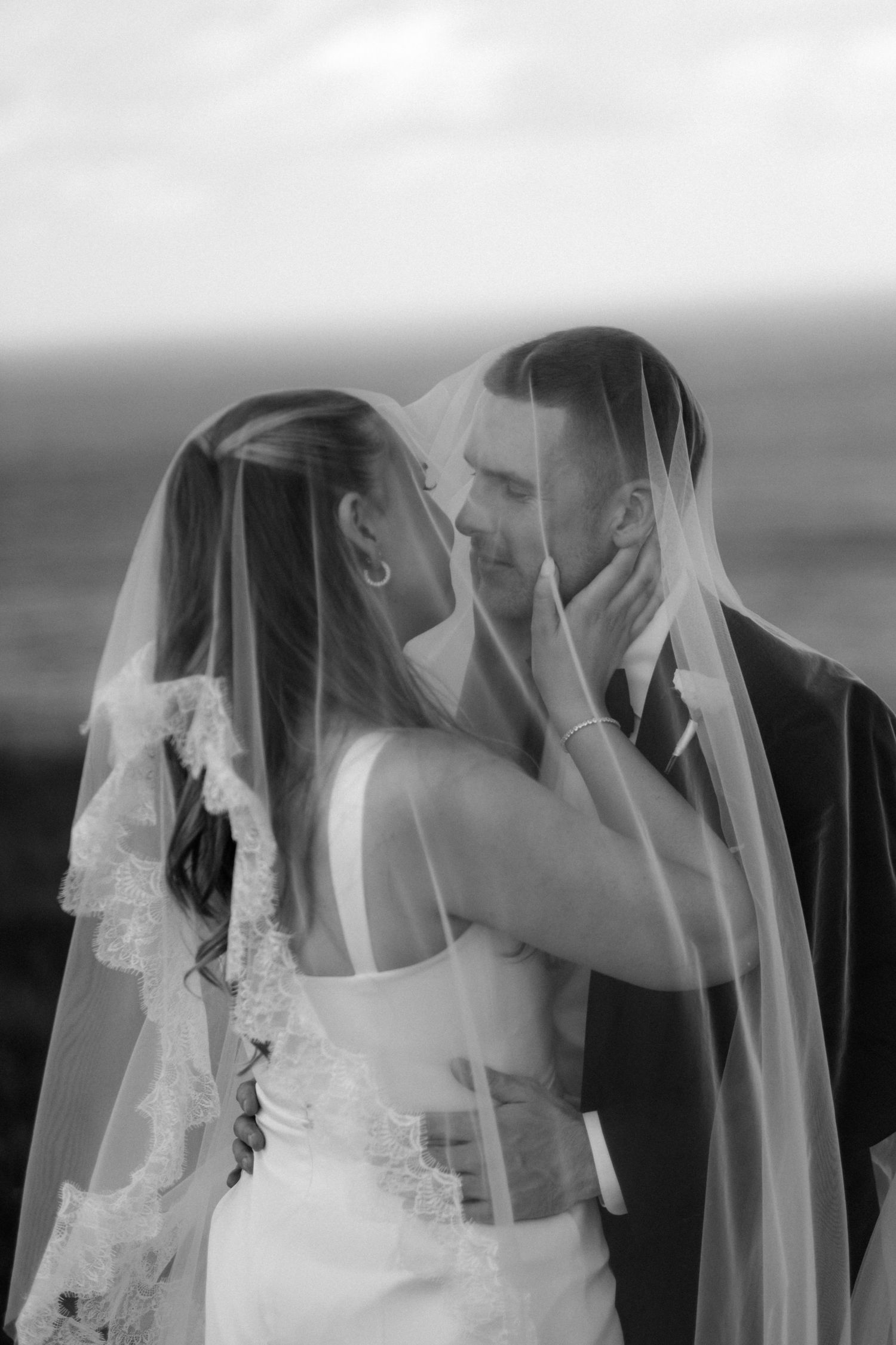 Black and white romantic photo of bride and groom sharing intimate moment under lace wedding veil.