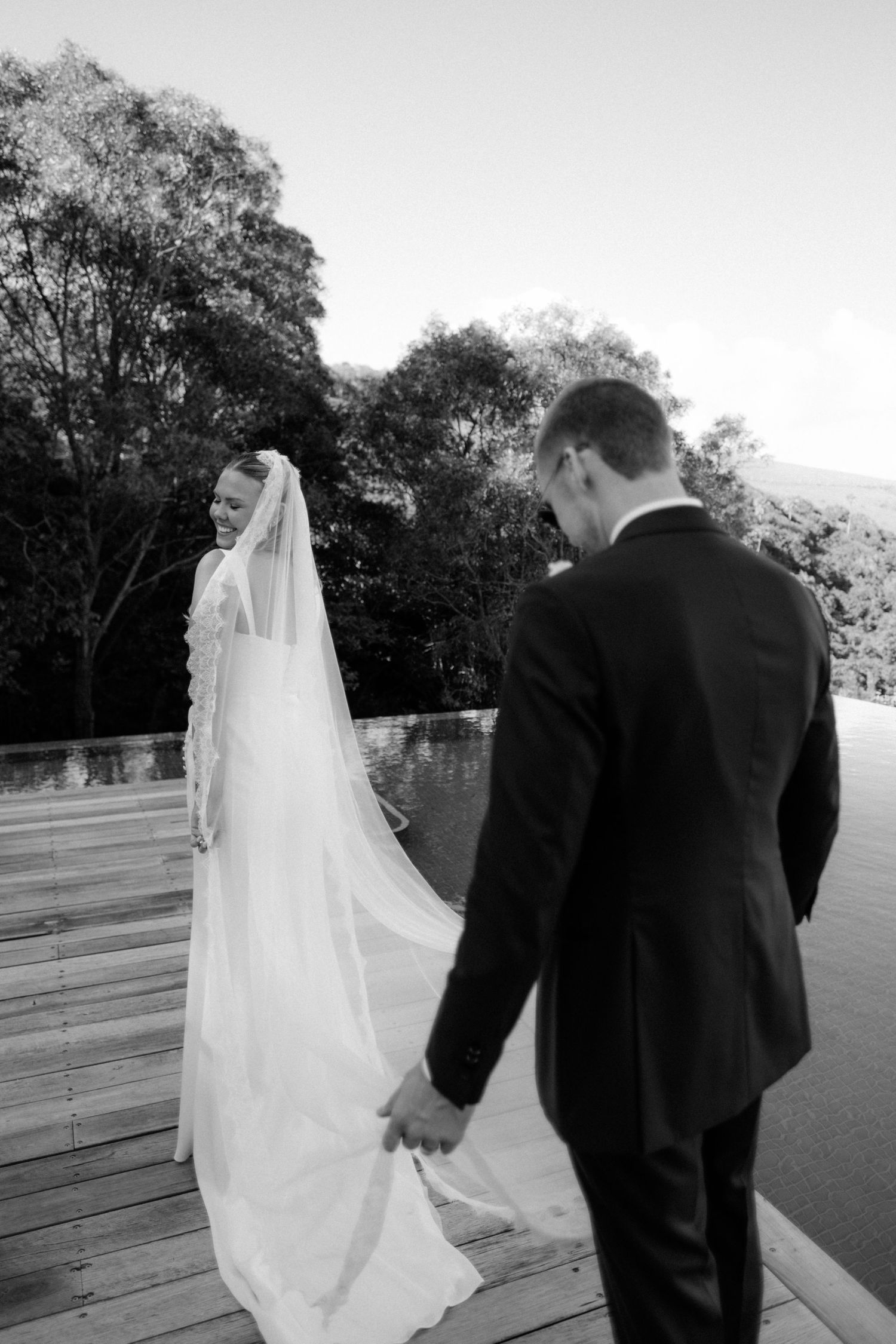 Bride and groom holding hands on a wooden deck surrounded by lush trees in a romantic black and white wedding photo.